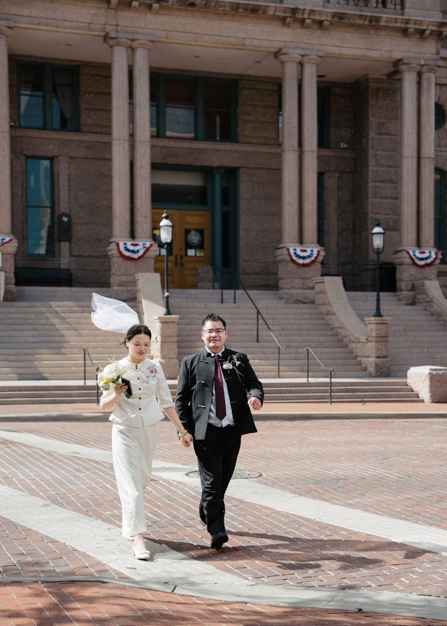 Newlywed couple walking hand in hand in front of a grand courthouse building on their wedding day.