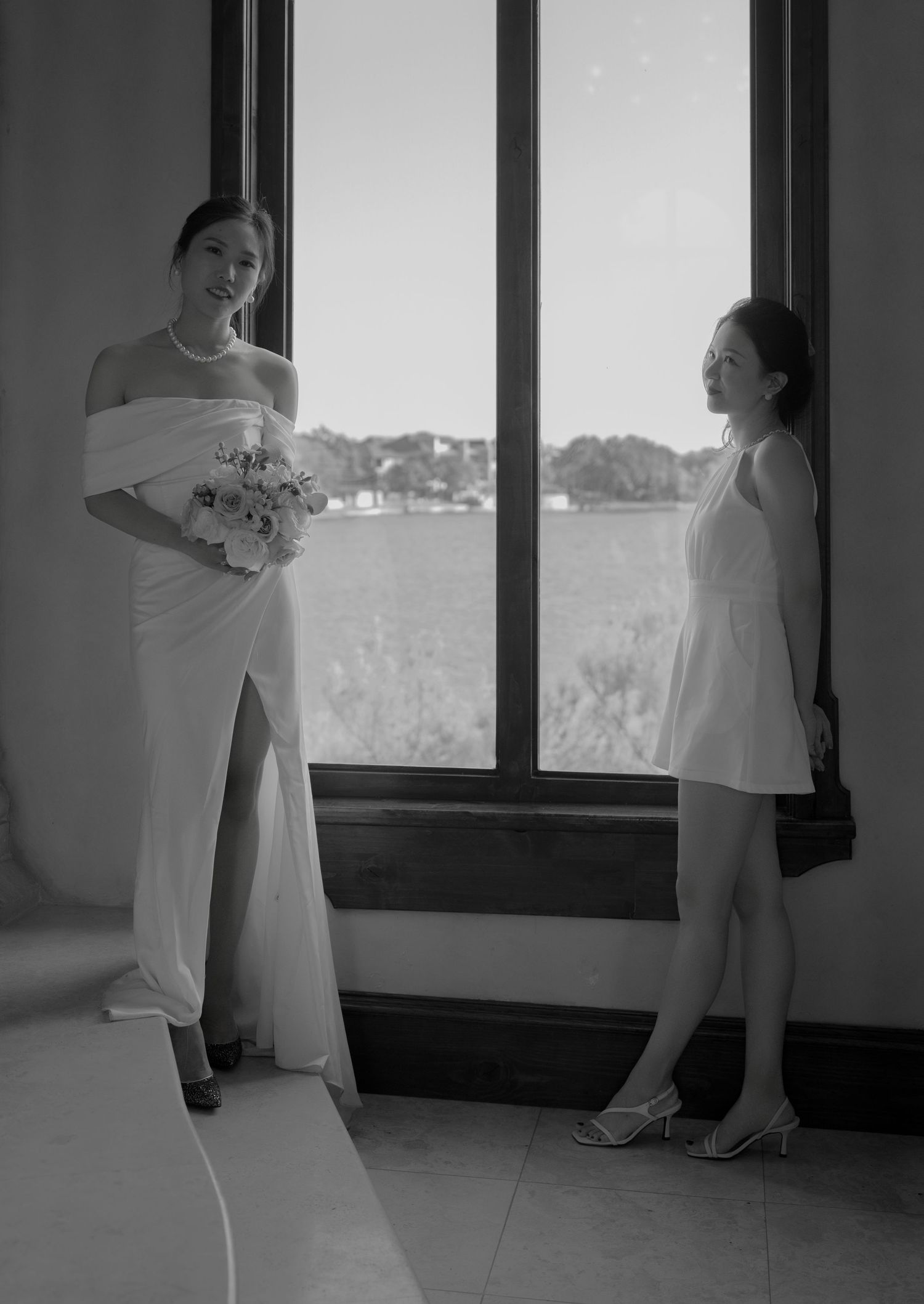 Bride in white off-shoulder gown holds bouquet while standing near a window with a guest in black and white photo.
