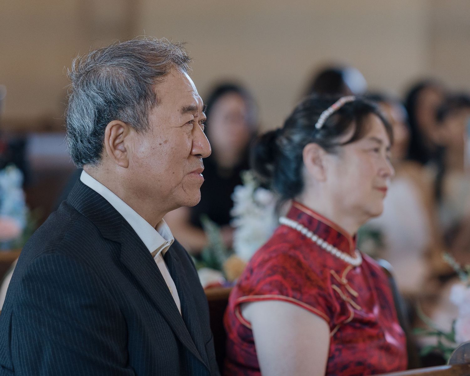Elderly Asian couple seated in church pews during a wedding ceremony, man in suit, woman in red qipao.