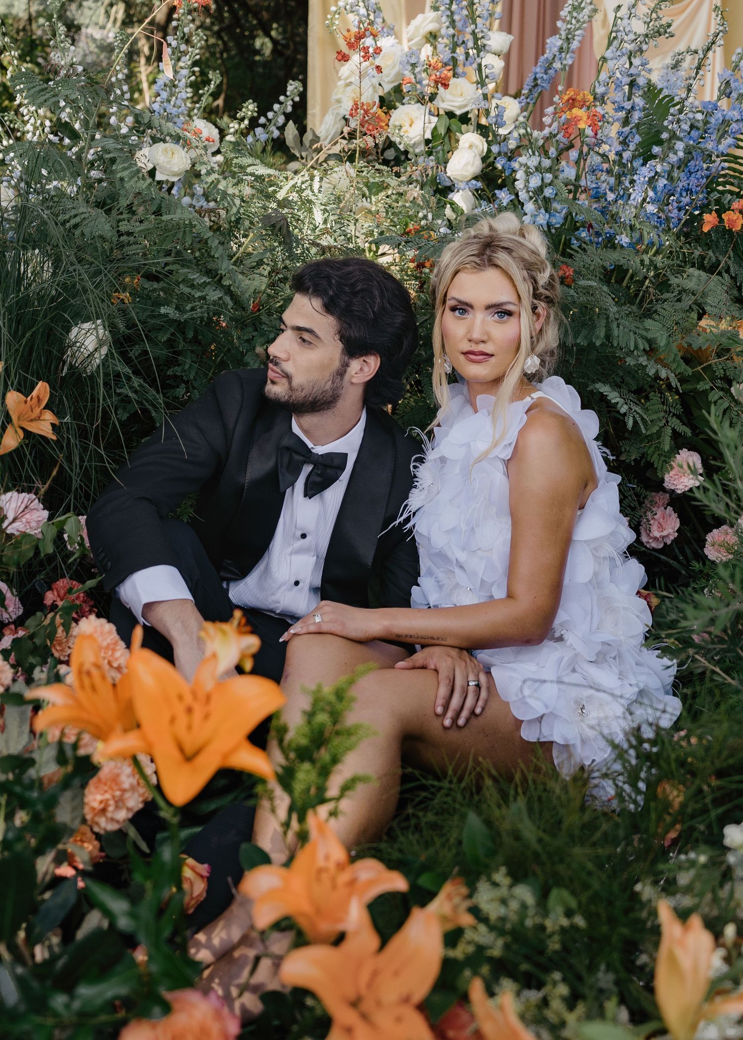 Couple sits surrounded by lush garden florals, bride in white feathered mini dress, groom in black tuxedo.