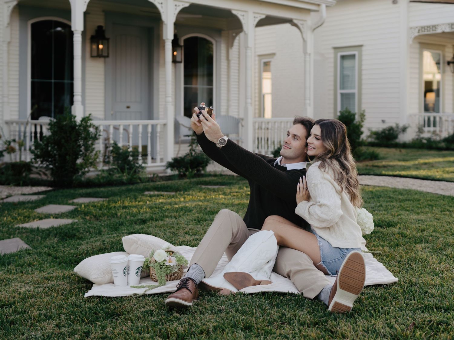 Engaged couple taking a selfie while sitting on a blanket on the lawn outside a white cottage.