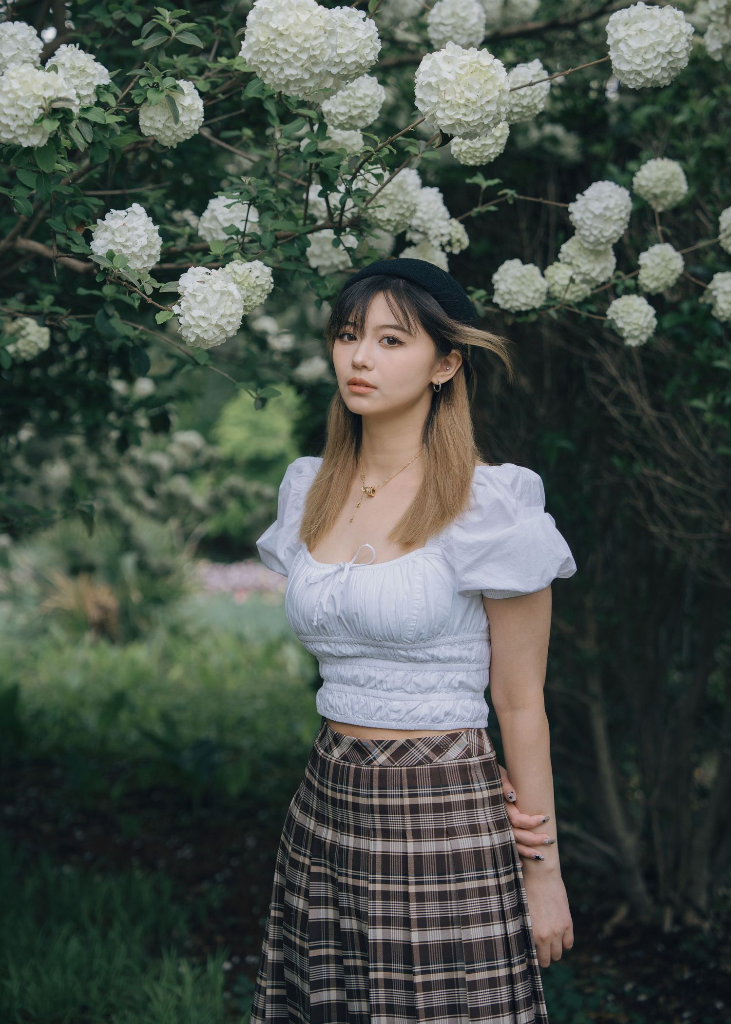 Young woman in white puff sleeve top and plaid skirt with black beret standing near white hydrangeas.