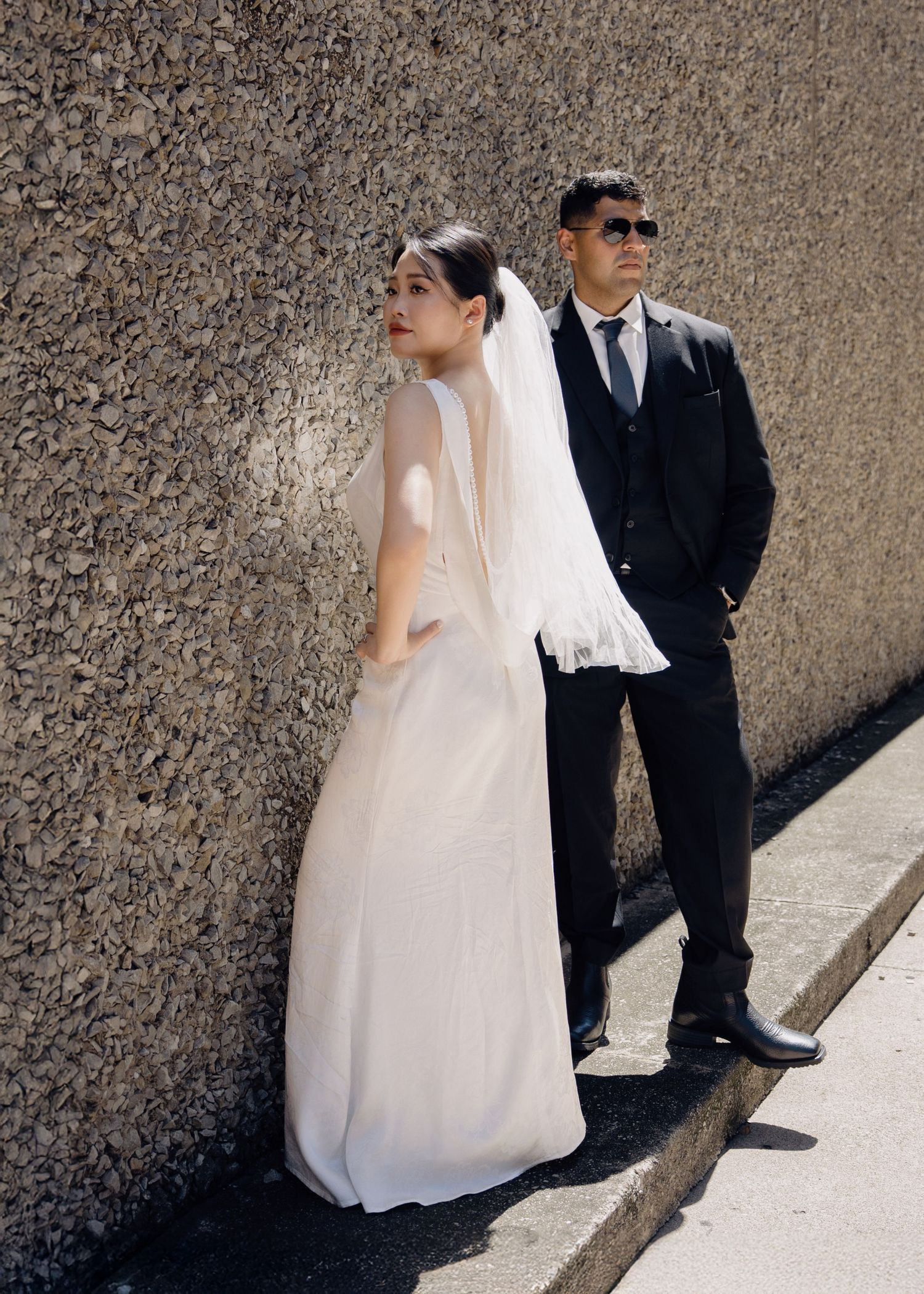 Bride and groom pose against a textured stone wall outdoors, she wears a flowing white gown with veil, he wears sunglasses.