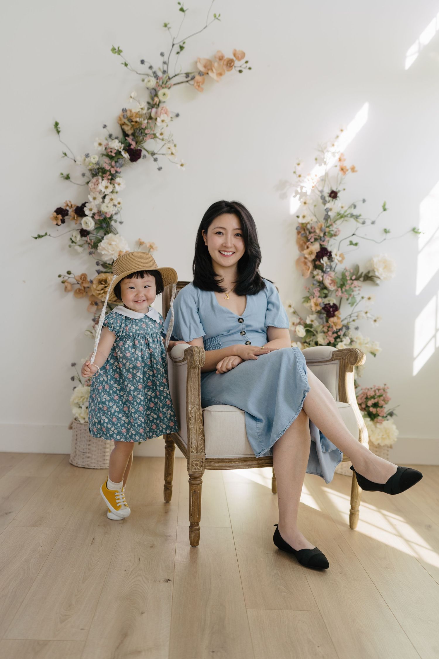 An Asian mother in a blue dress sits on a vintage chair beside her toddler daughter in a floral dress.
