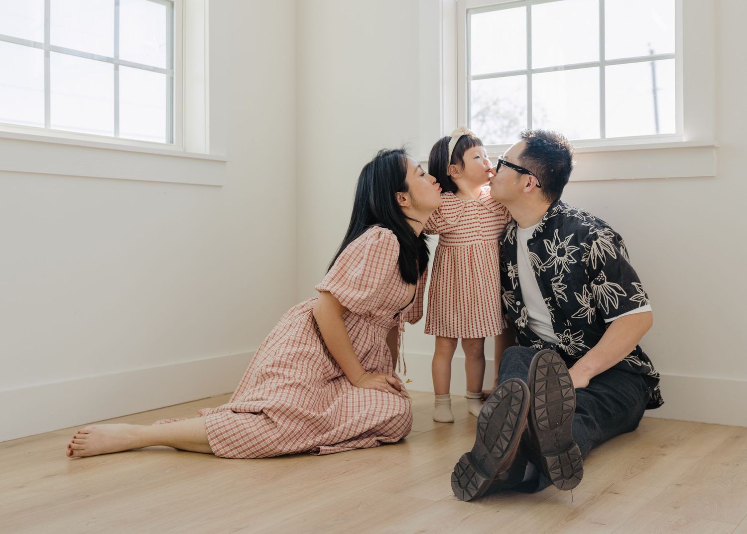 An Asian couple leans in to kiss their smiling toddler daughter between them on a bright hardwood floor.