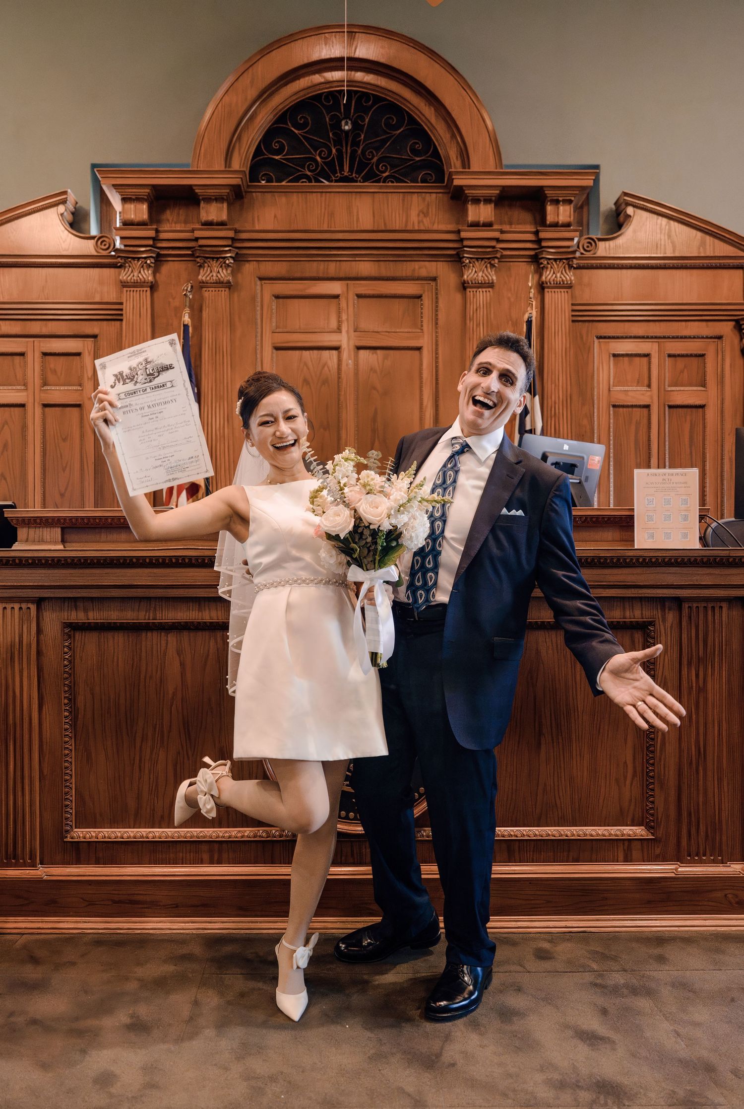 Ecstatic newlyweds celebrate holding marriage certificate and bouquet in front of courthouse bench.