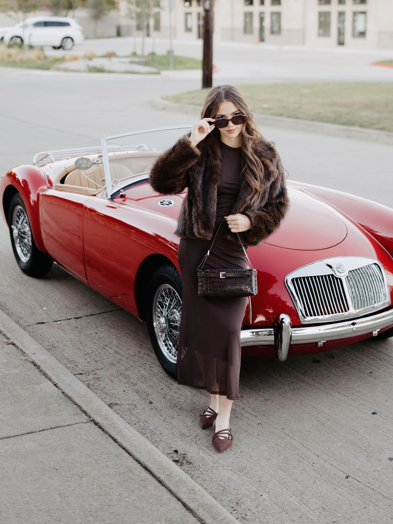 Woman in fur coat and brown dress posing beside a classic red MG convertible sports car.