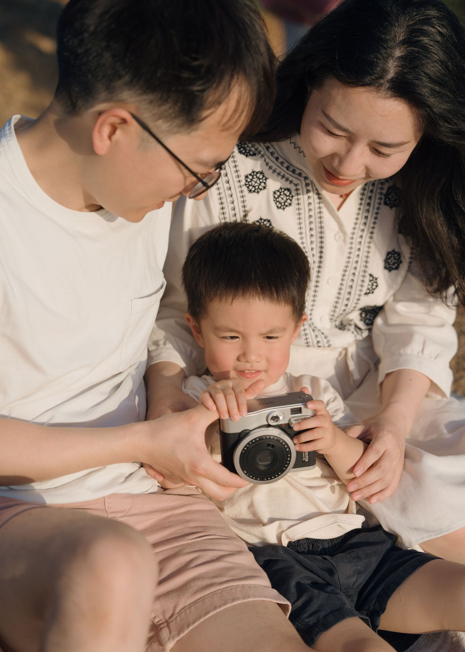 Parents and toddler son sit together outdoors as child examines a vintage film camera with curiosity.