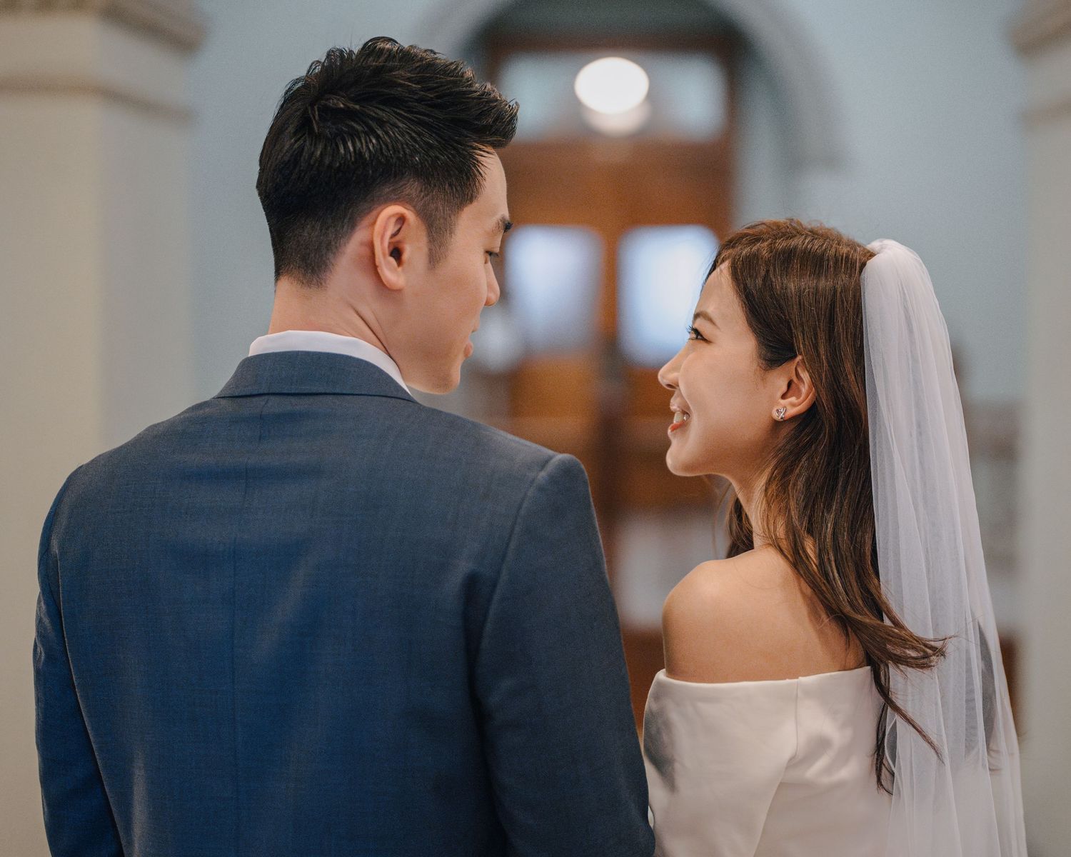 Asian bride in white off-shoulder gown smiling at groom in blue suit inside elegant venue.