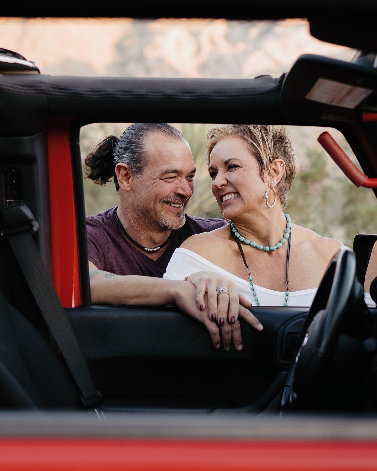 Happy couple smiling at each other inside a red open-top Jeep during golden hour outdoors.