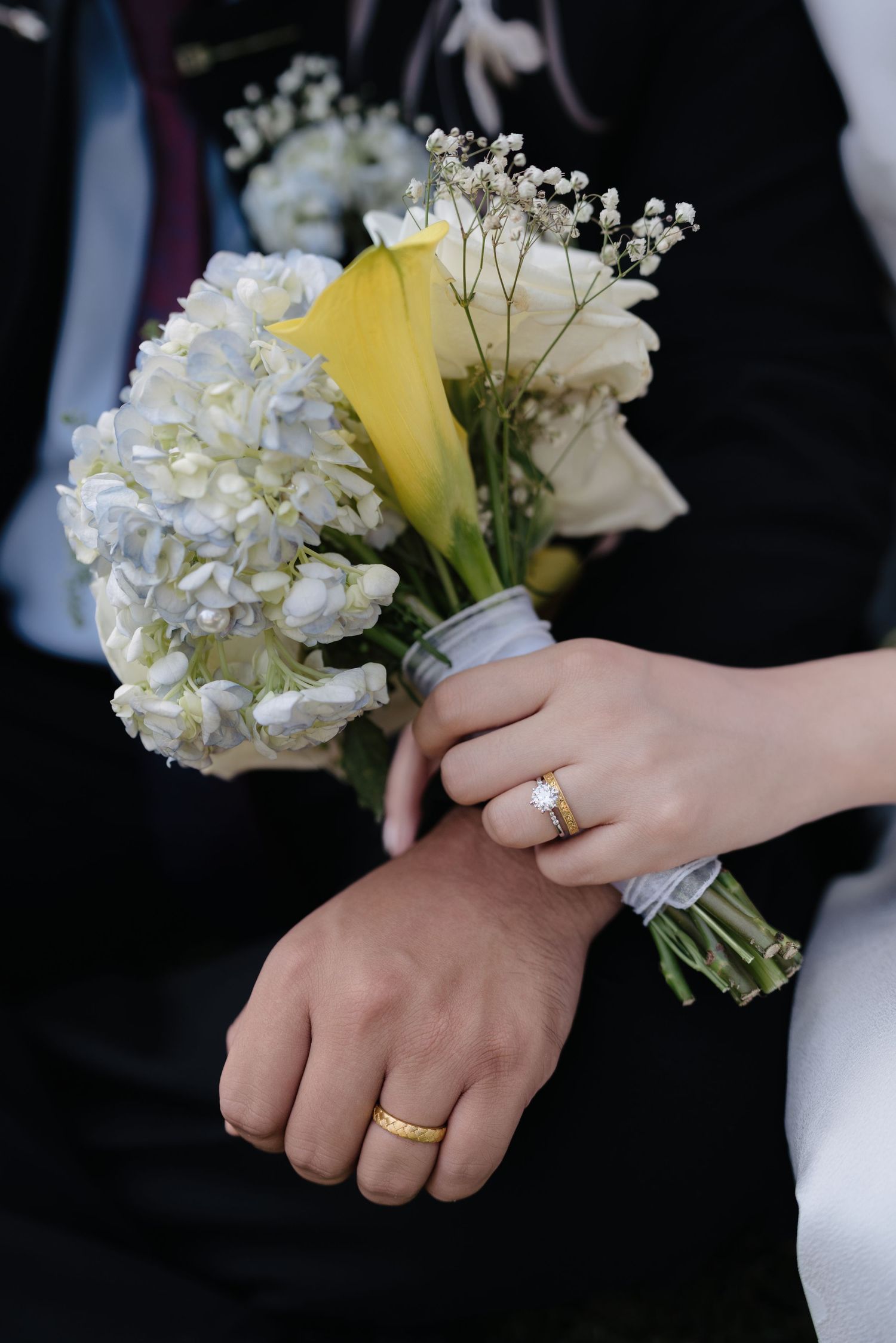 Bride and groom holding a white and yellow wedding bouquet, showing gold wedding bands on their hands.