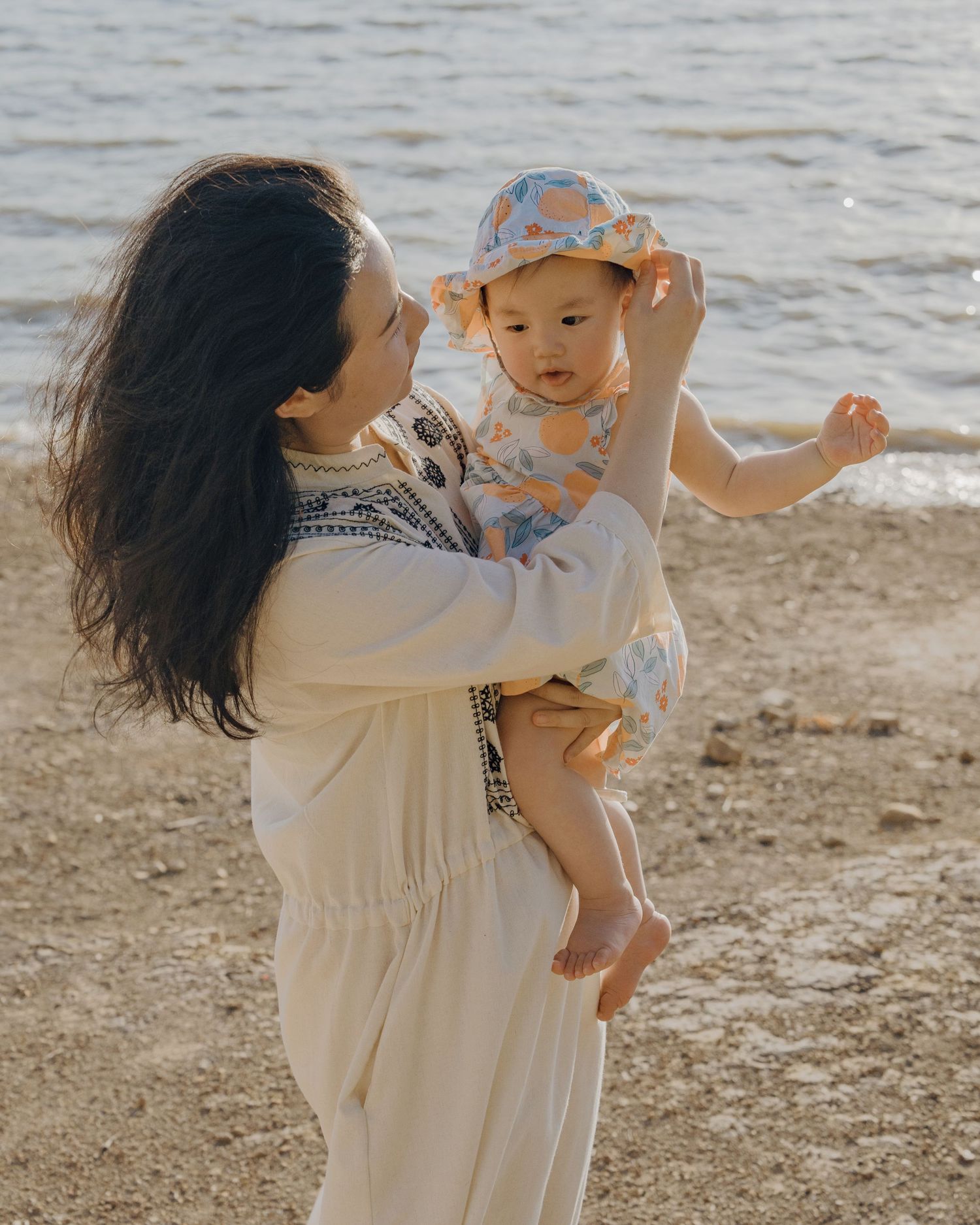 Mother in white embroidered dress holds baby wearing a floral bucket hat on a sandy beach.