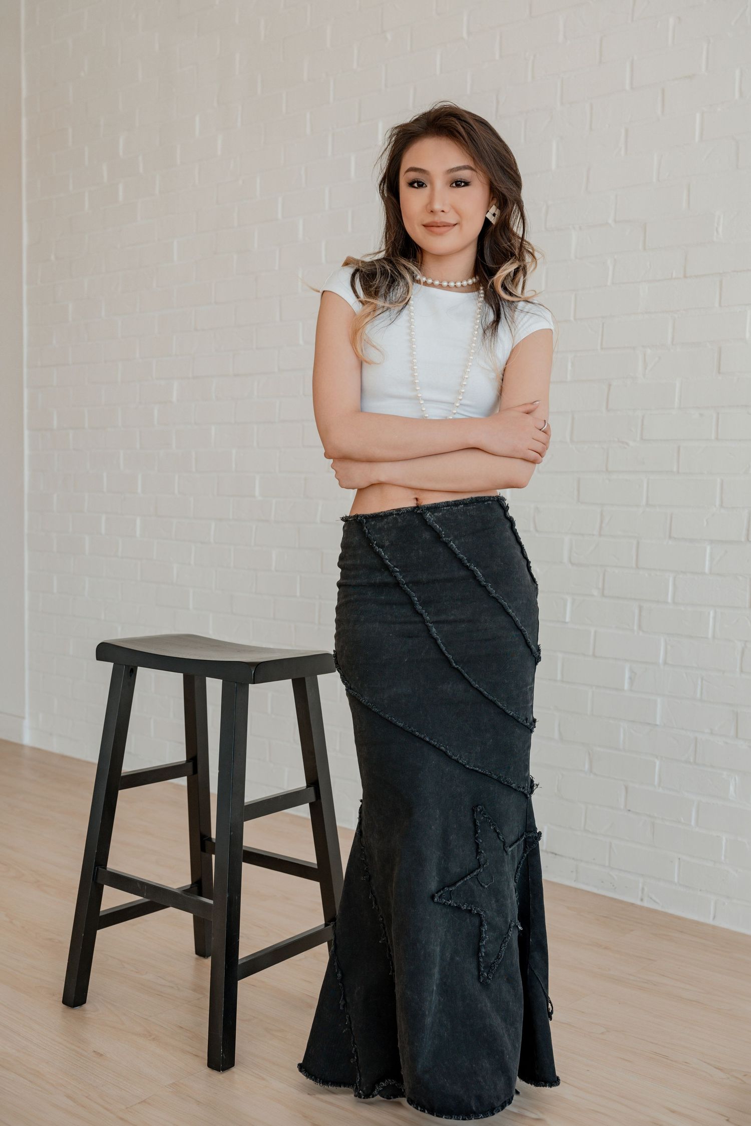 Confident young woman in white crop top and dark denim maxi skirt posing by wooden stool.