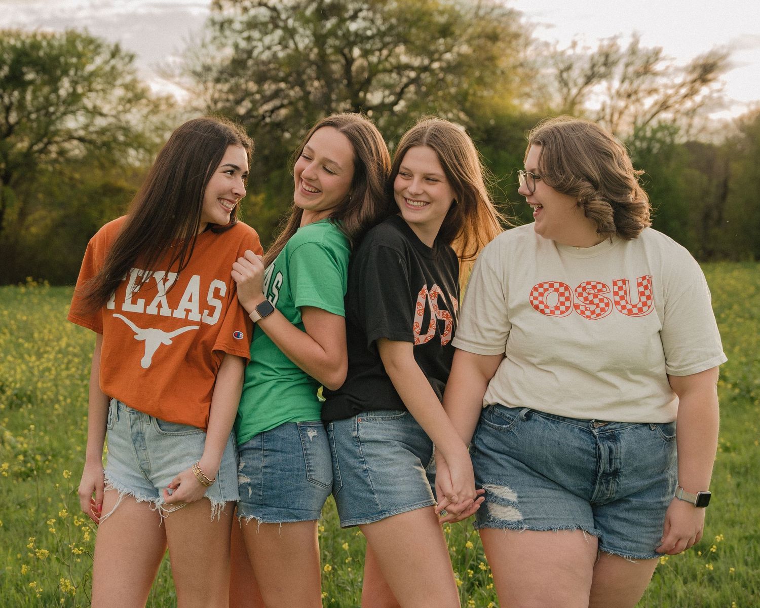 Four girls laughing together in a sunny field wearing Texas, OSU, and college graphic tees.