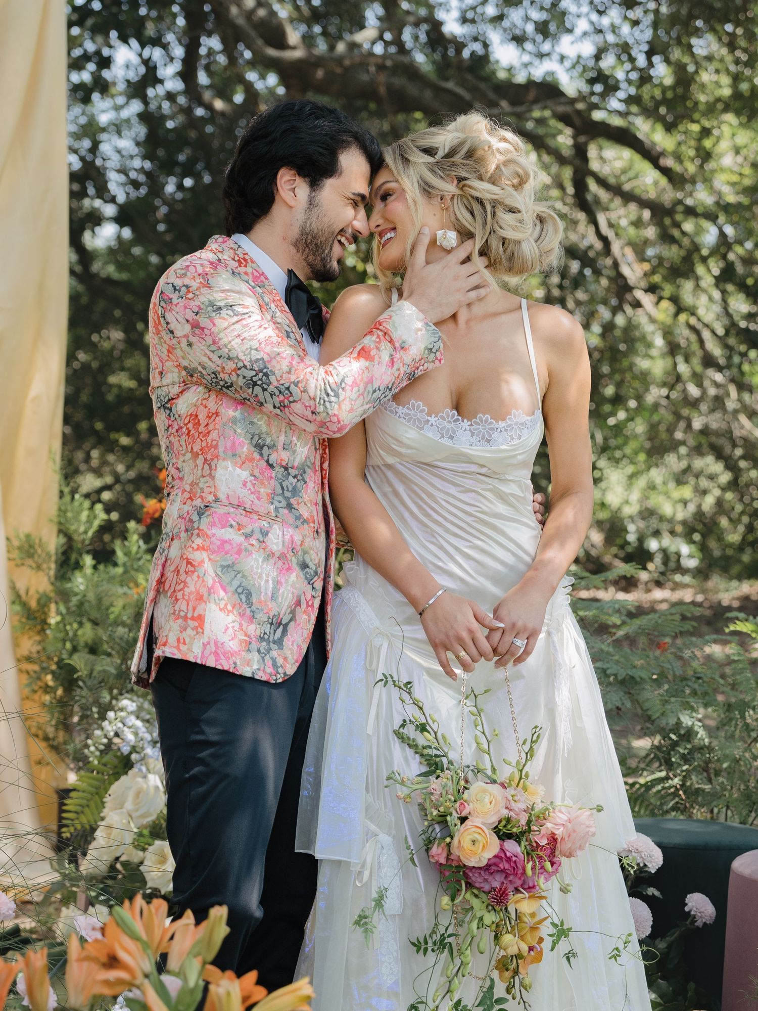 Smiling couple shares romantic moment at outdoor wedding, bride holding colorful bouquet, groom in floral blazer.
