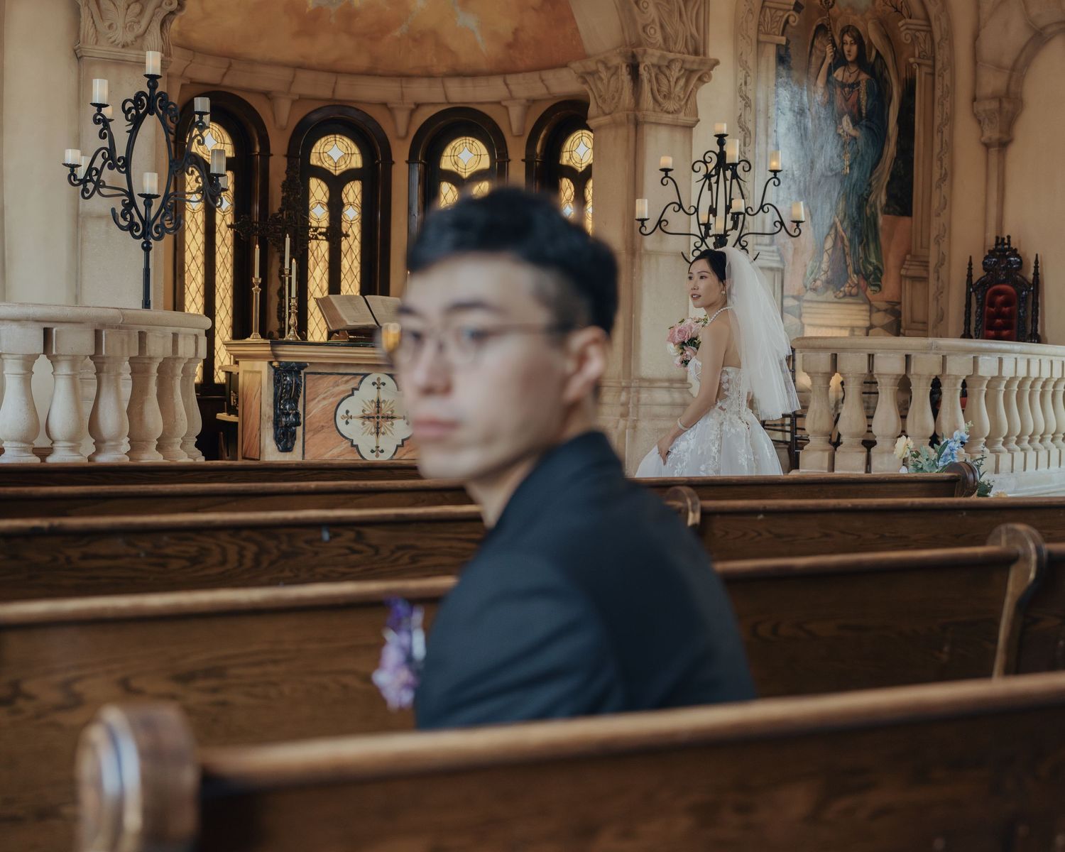 Groom in dark suit sits in church pew as bride in white gown stands at altar in background.