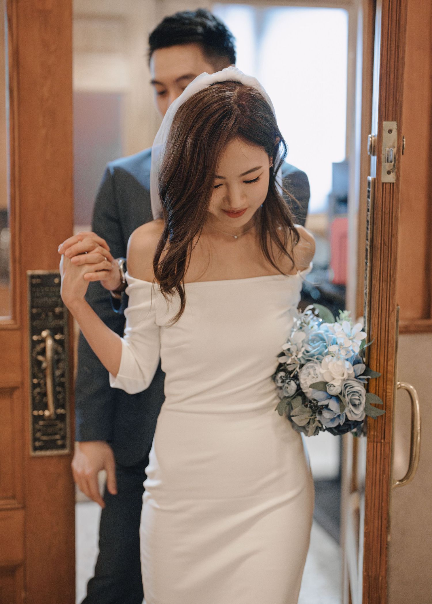 Beautiful Asian bride in fitted white off-shoulder wedding dress holding blue bouquet at wooden doors.