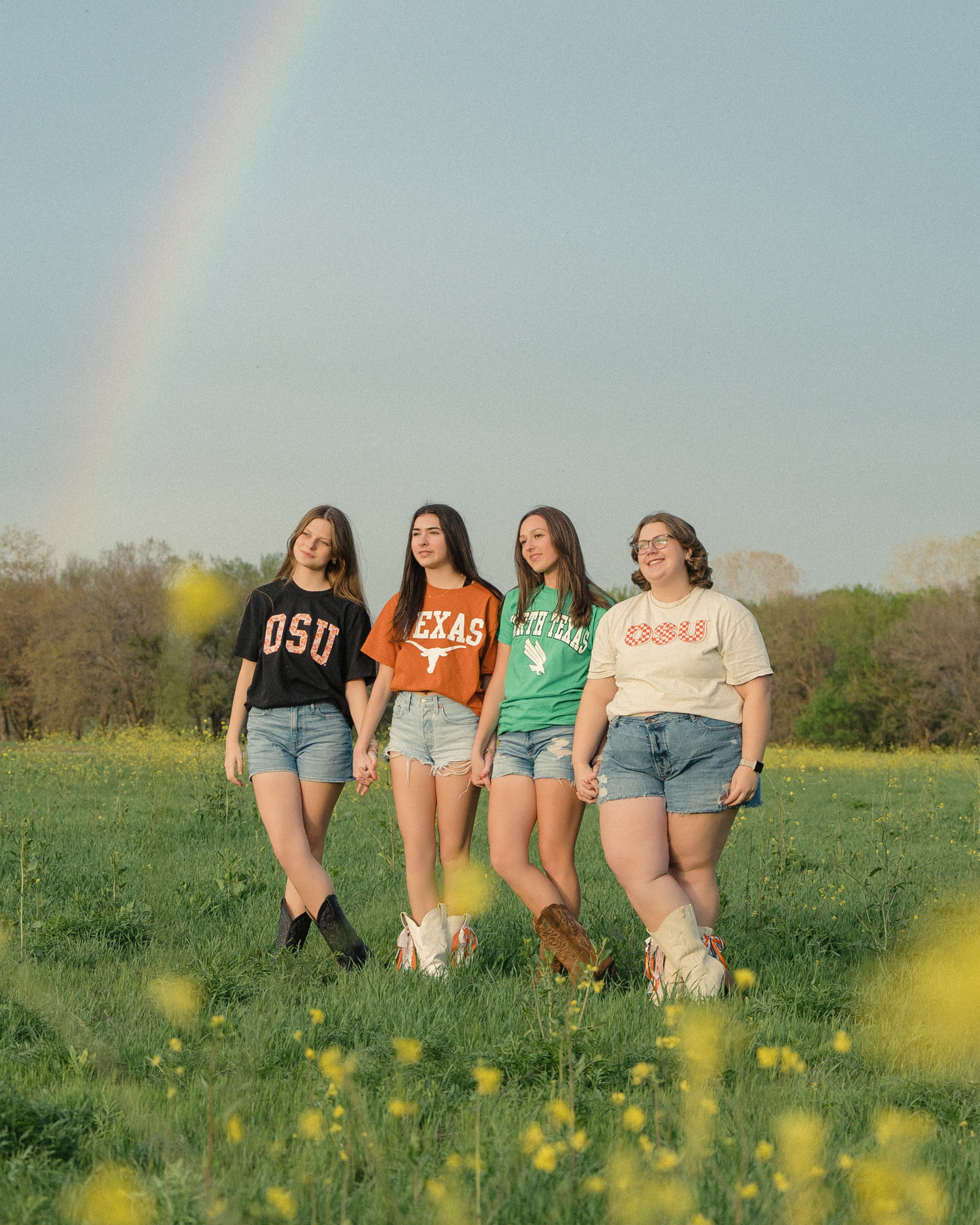 Four girls posing in a green field with a rainbow behind them wearing OSU, Texas, and college tees.