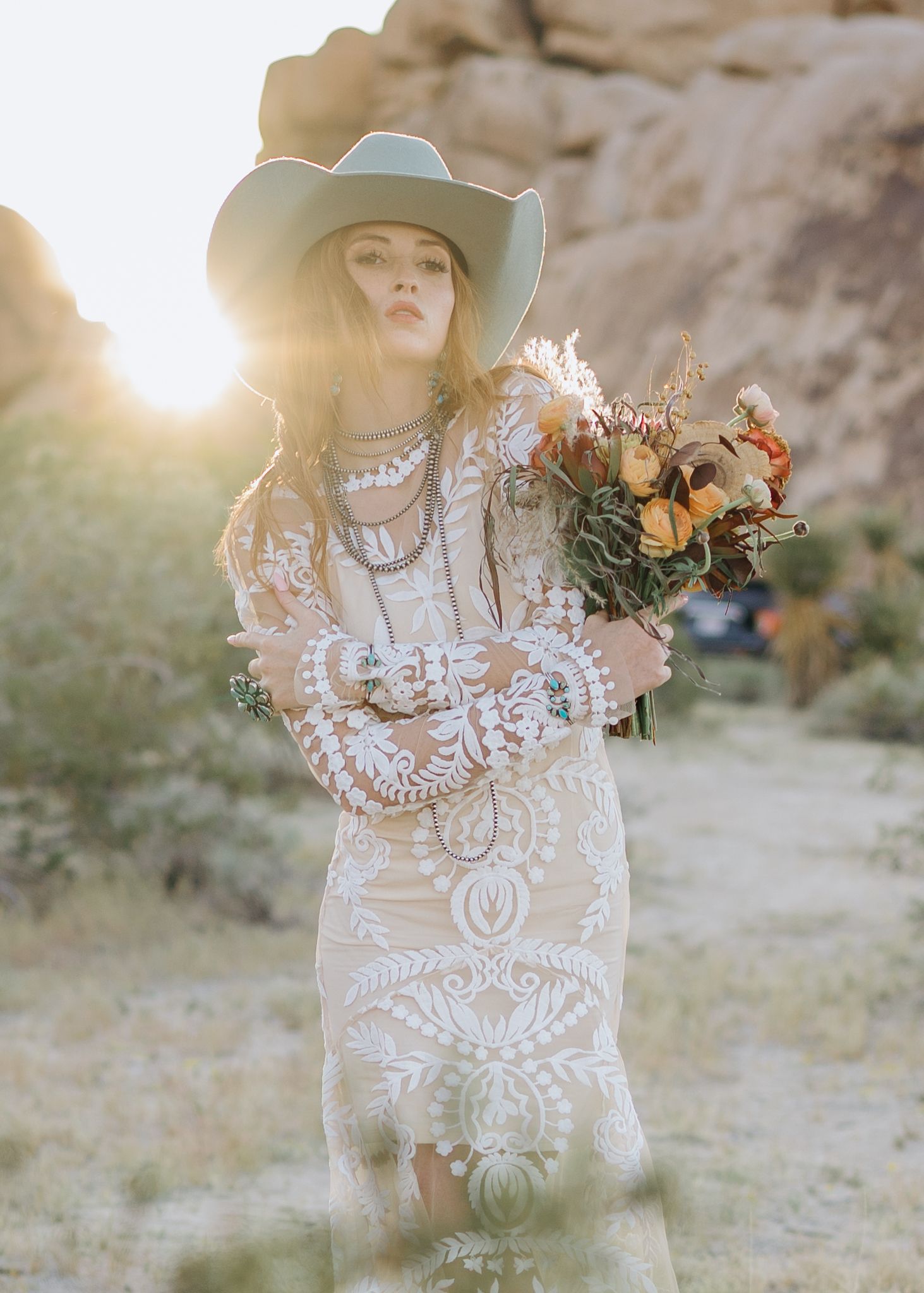 Boho bride in lace dress and cowboy hat holding colorful bouquet in desert landscape at golden hour.