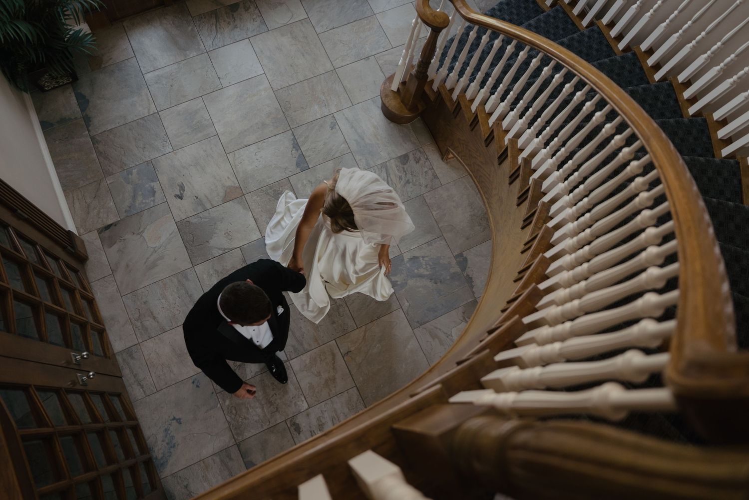 Aerial view of bride and groom walking on tiled floor beside elegant curved staircase.