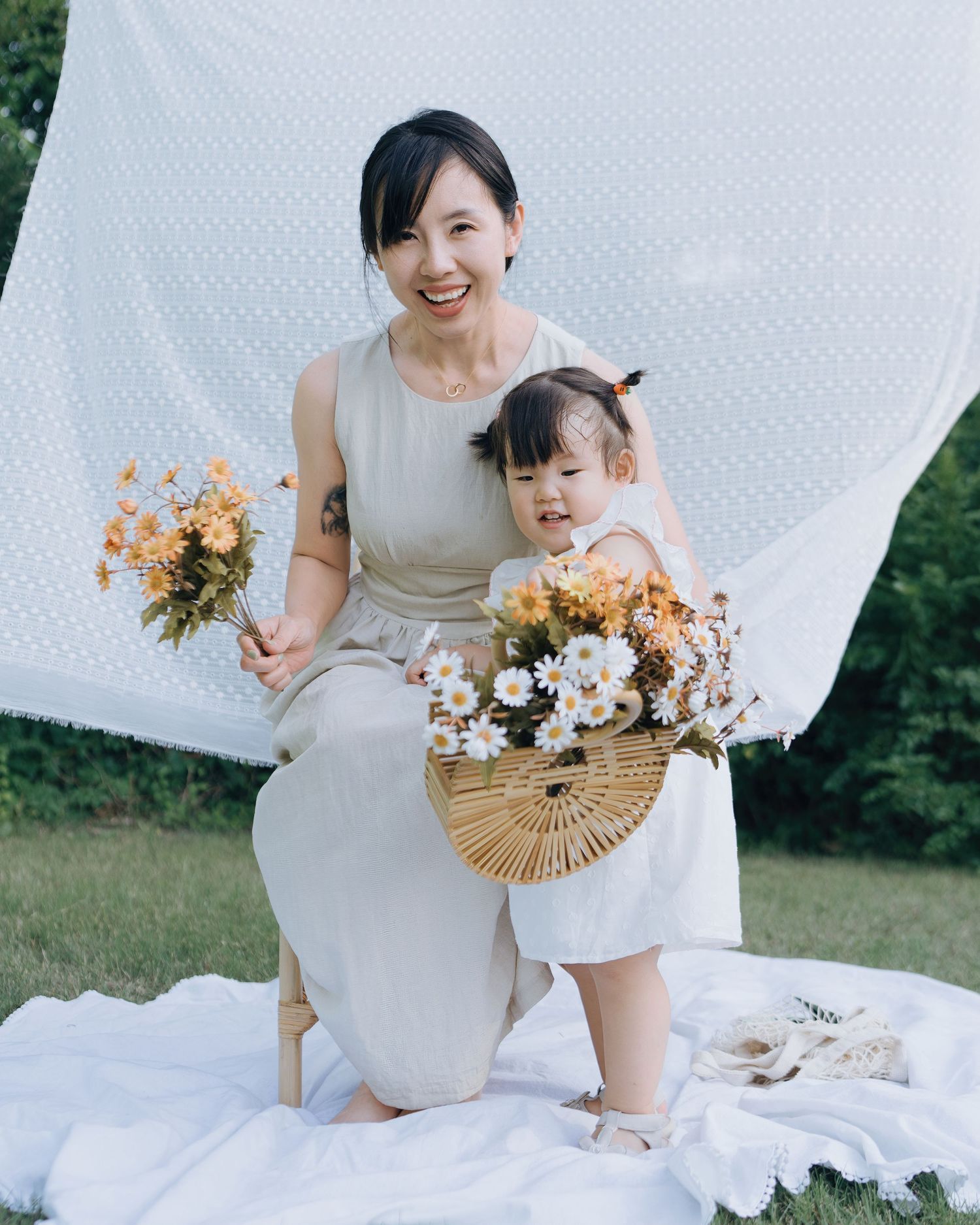 A smiling Asian mother and toddler daughter pose with yellow and white flowers in a sunny garden.