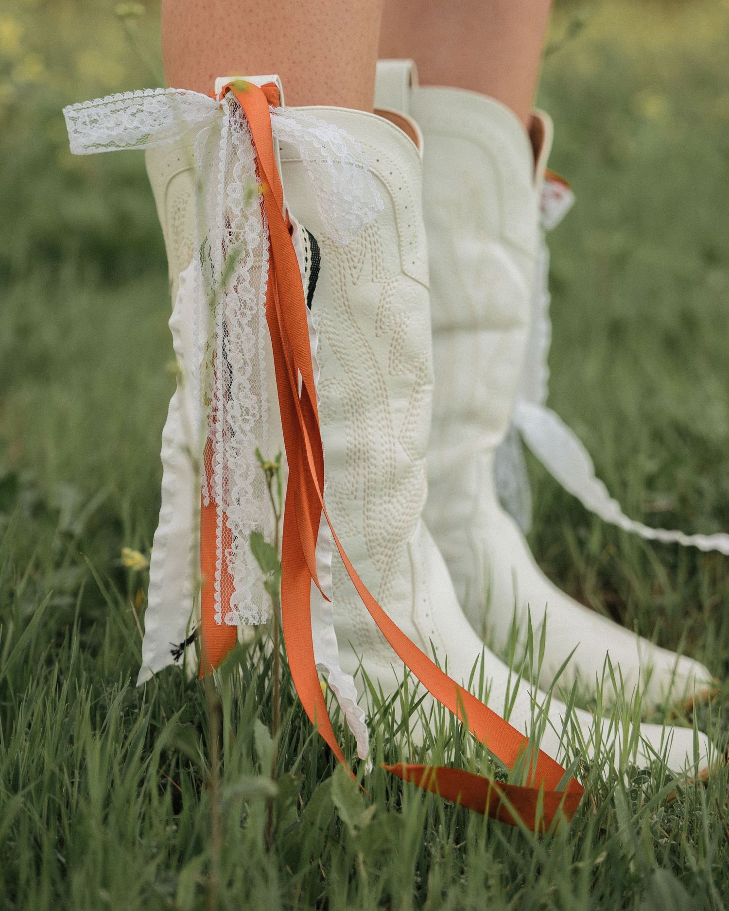 Back view of cream cowboy boots with orange ribbon and lace trim standing in green grass.