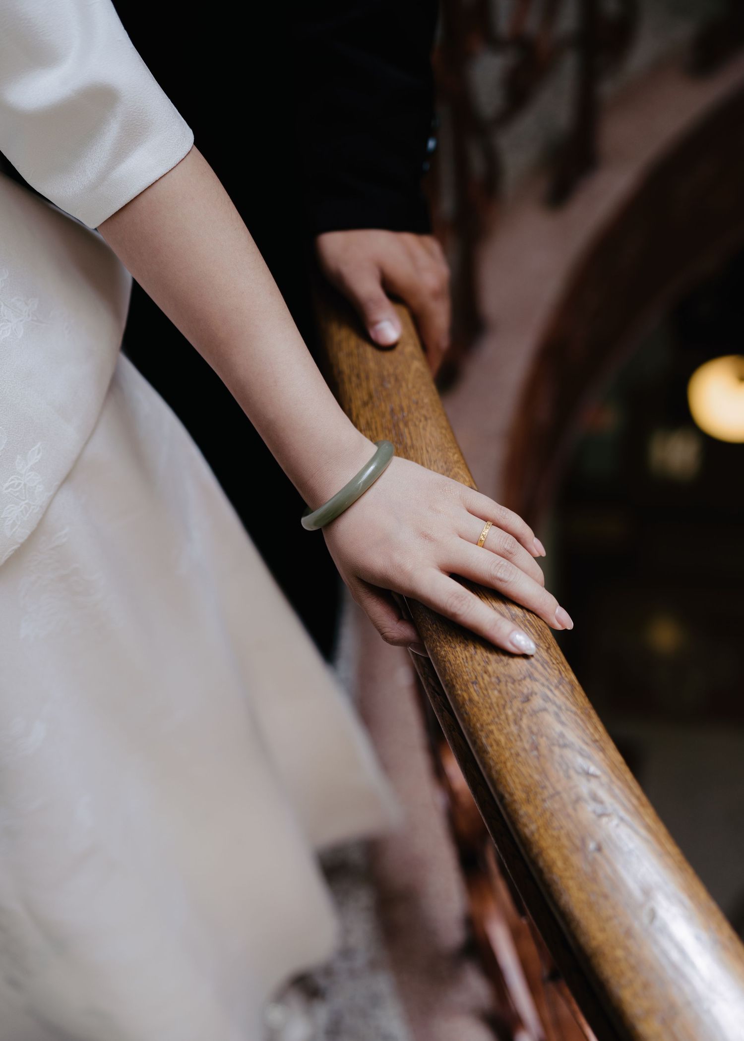 Bride and groom hands resting on wooden staircase railing, bride wearing gold ring and green bracelet.