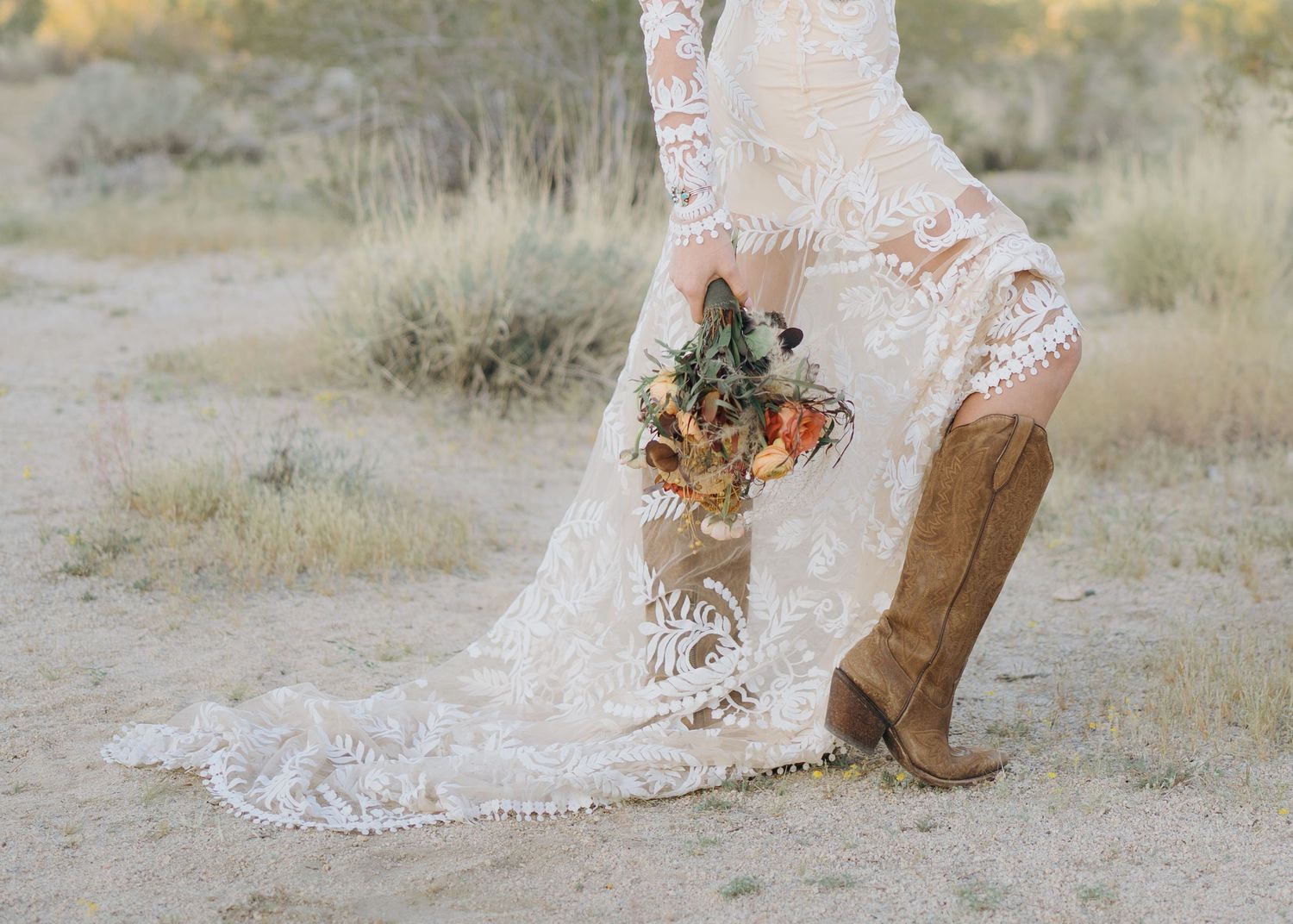 Close-up of bride in lace wedding dress and cowboy boots holding colorful wildflower bouquet.