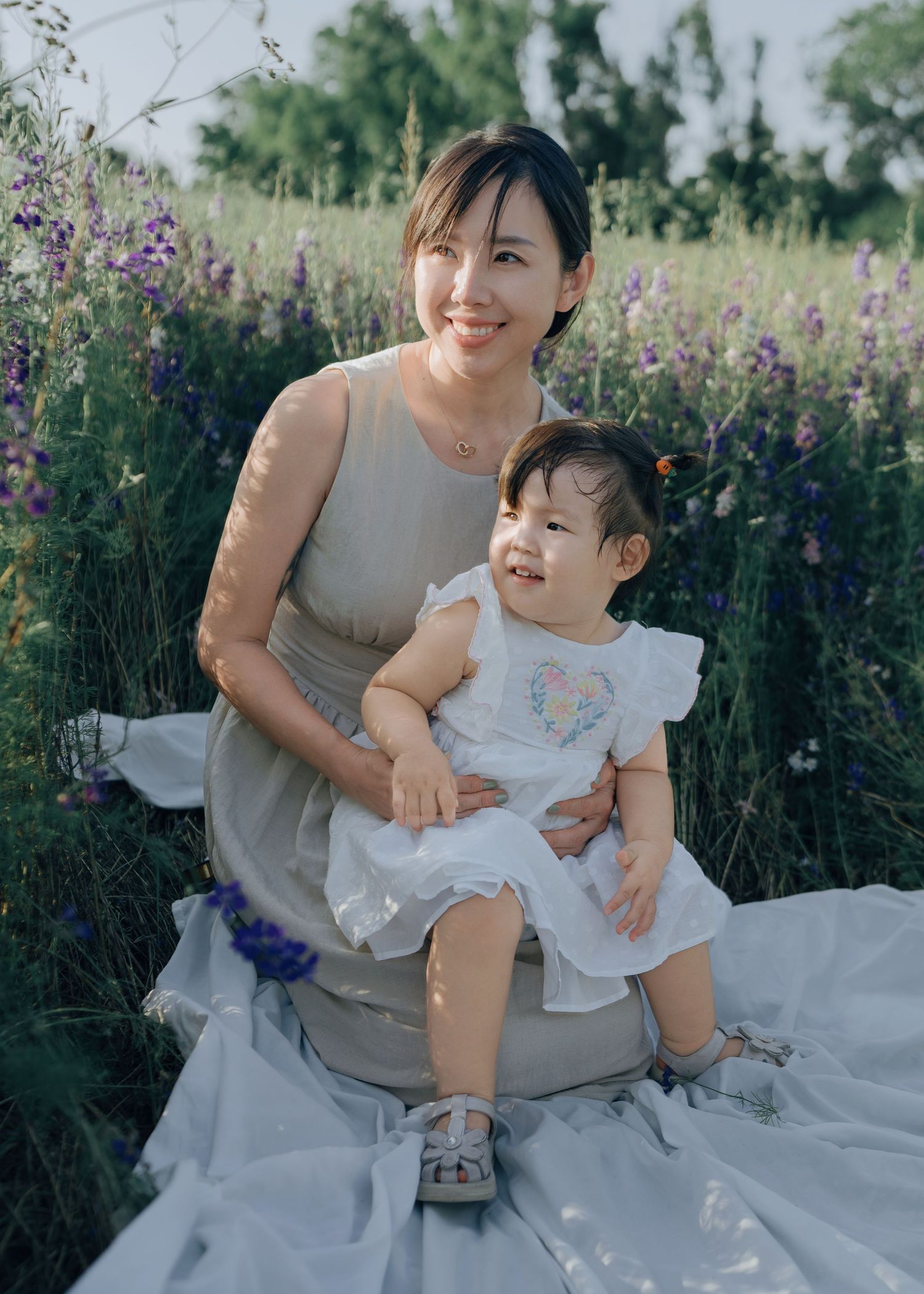 Smiling Asian mother holding cheerful toddler daughter sitting in blooming purple flower field.