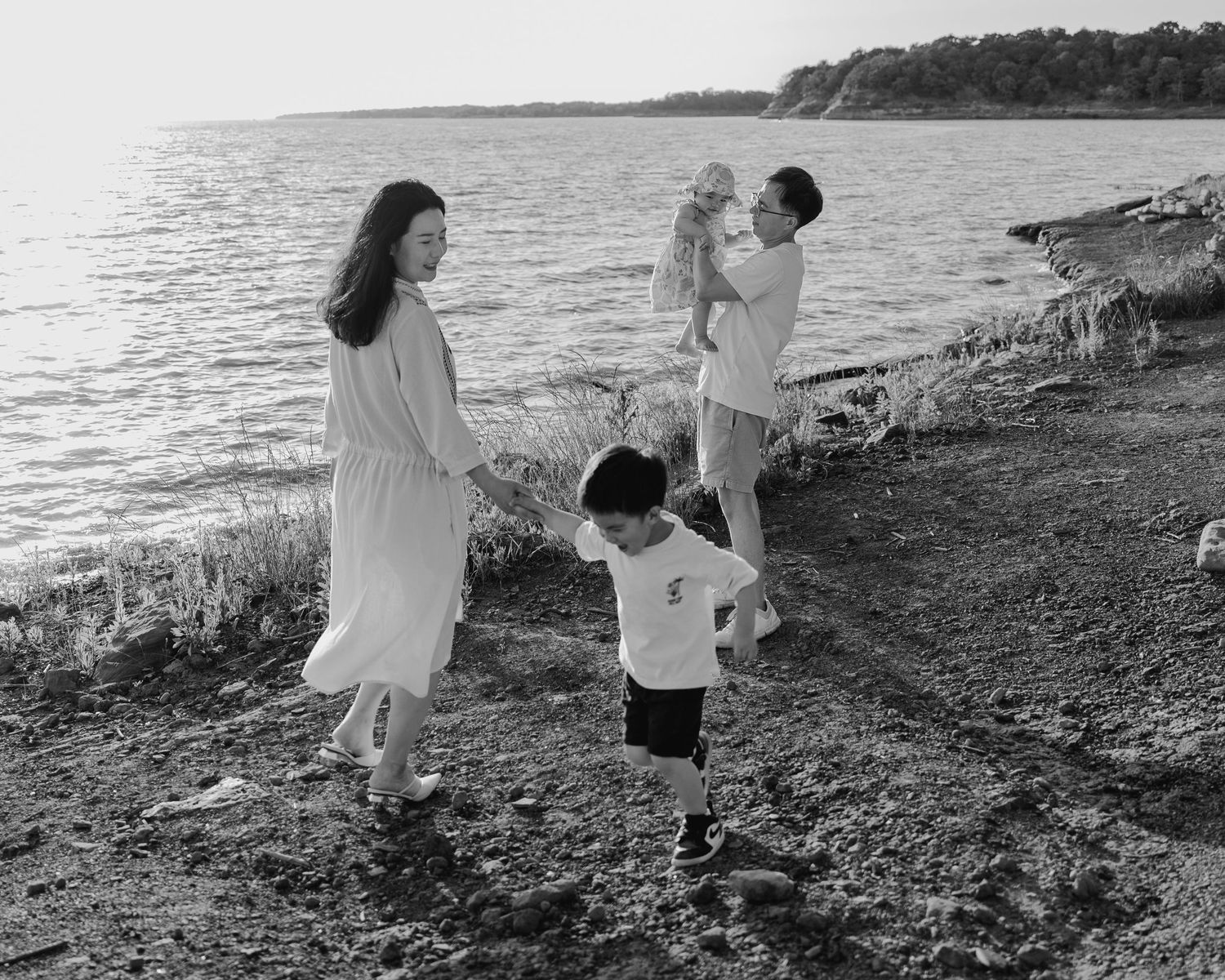 Black and white photo of a young Asian family walking along a rocky shoreline at sunset.