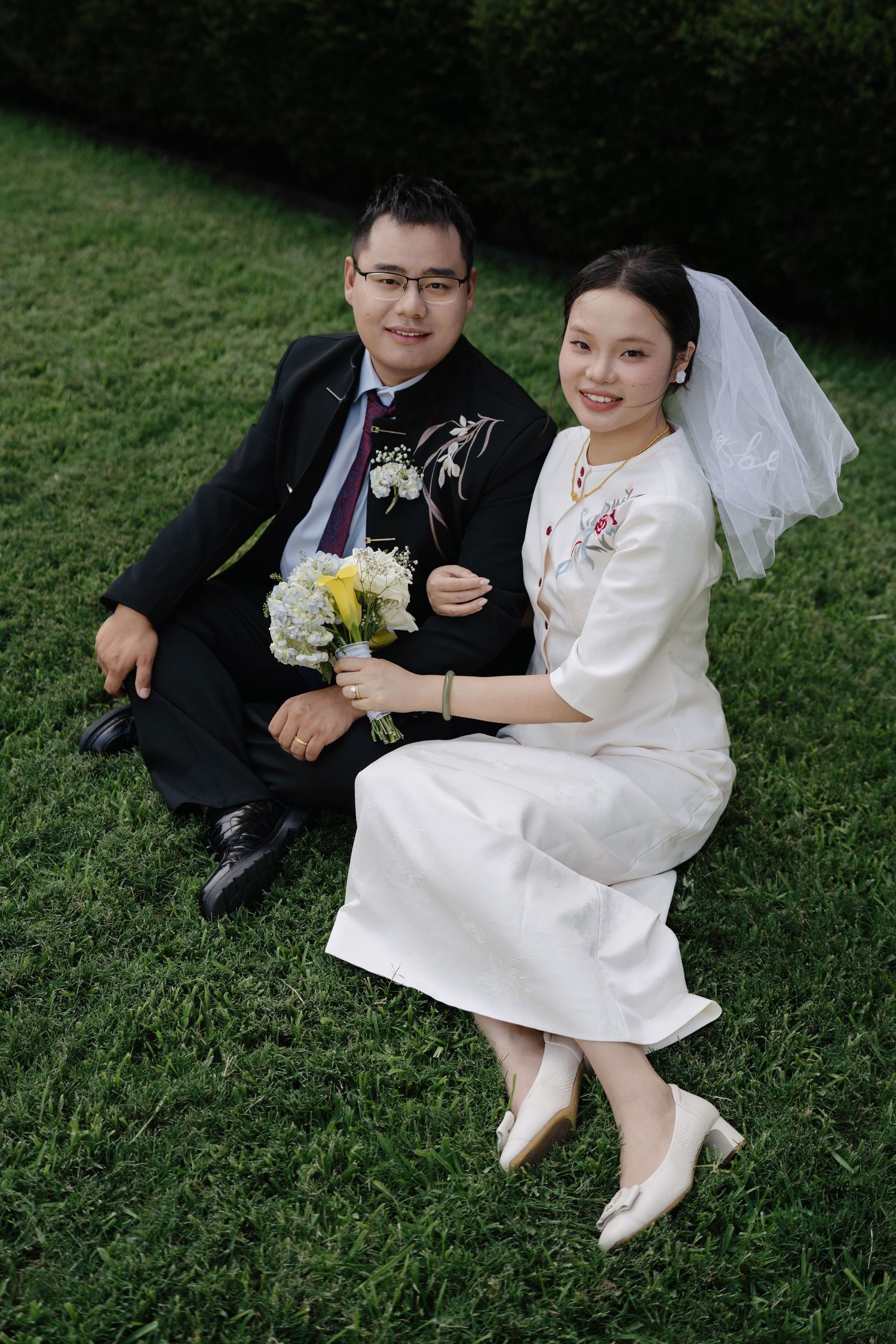 Asian newlyweds sitting on green grass, bride in white dress with veil holding bouquet, groom in black suit with glasses.