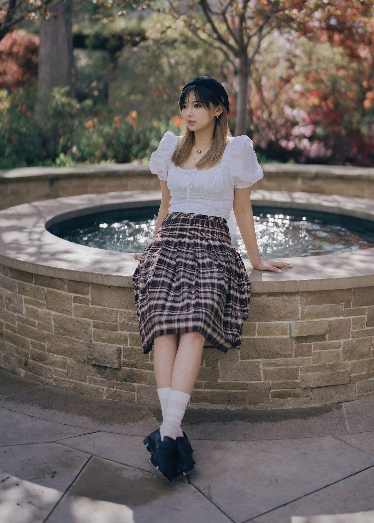 Young woman in plaid skirt and white puff-sleeve top leans against stone garden fountain.