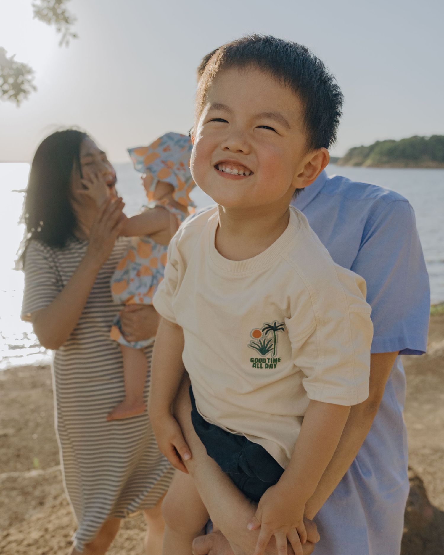 A joyful Asian boy smiling brightly outdoors near a lake while being held by his father.