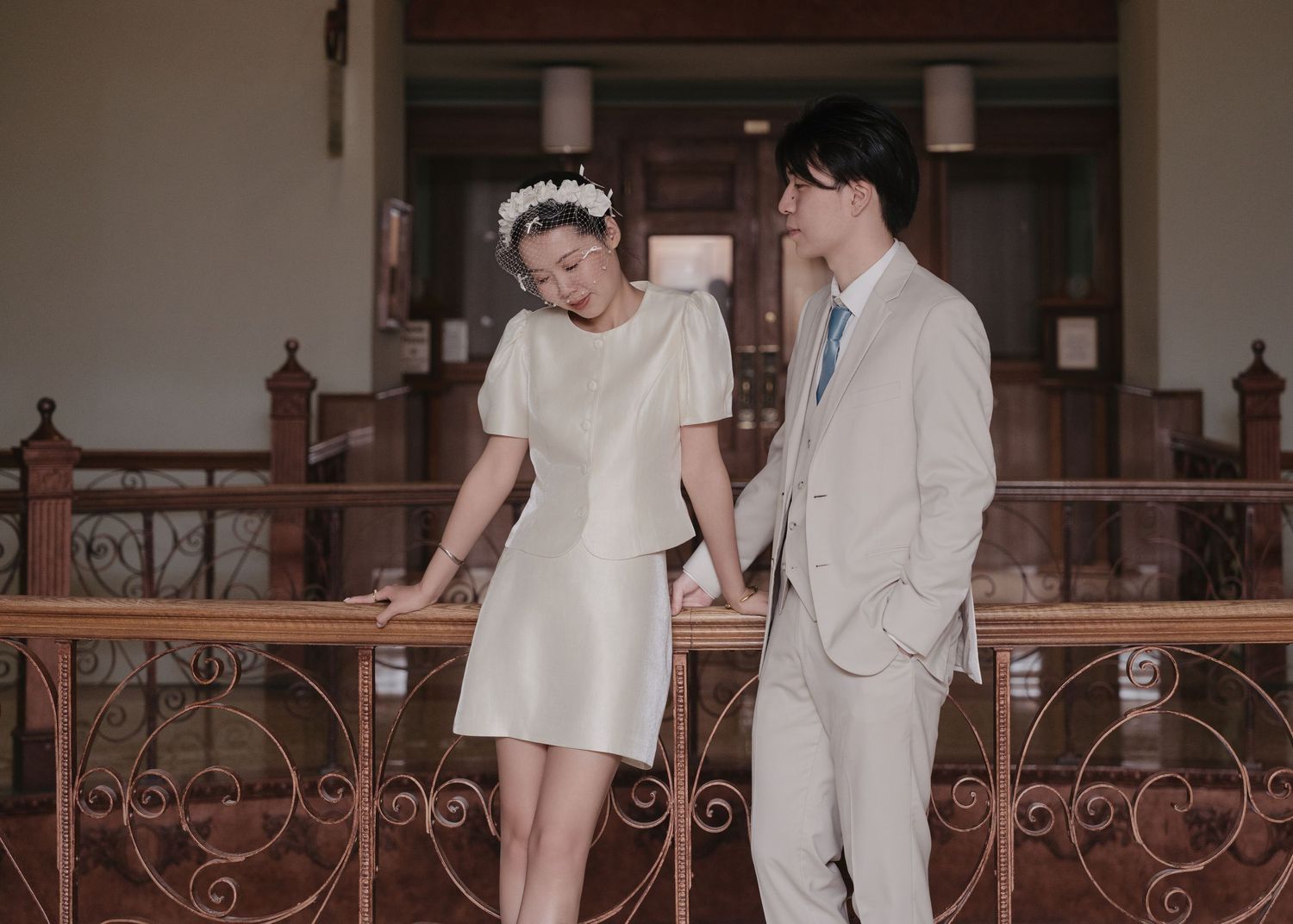 Elegant couple in wedding attire holds hands while leaning against an ornate iron railing indoors.