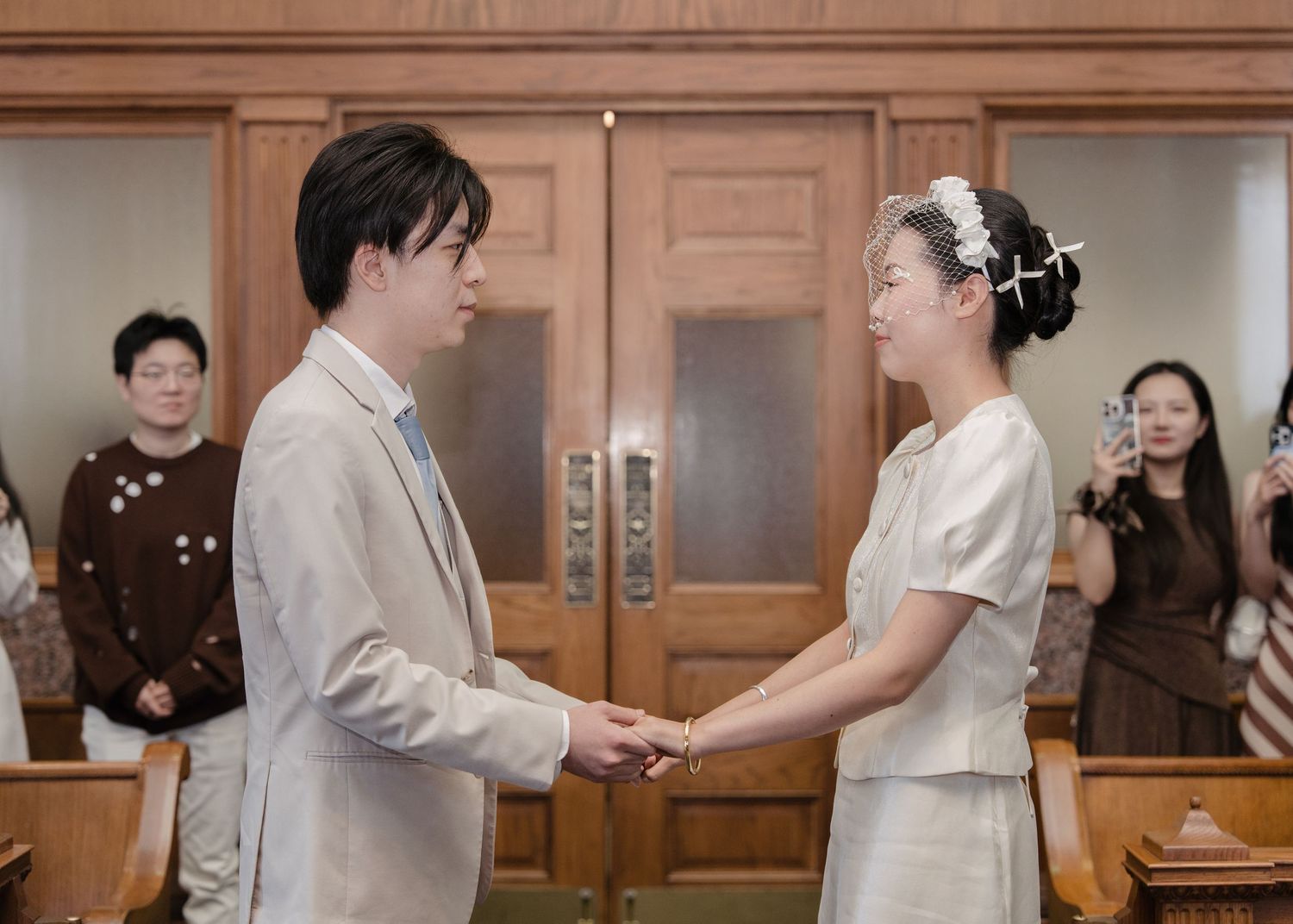 A couple exchanges vows holding hands at a civil wedding ceremony in a wood-paneled room with guests watching.