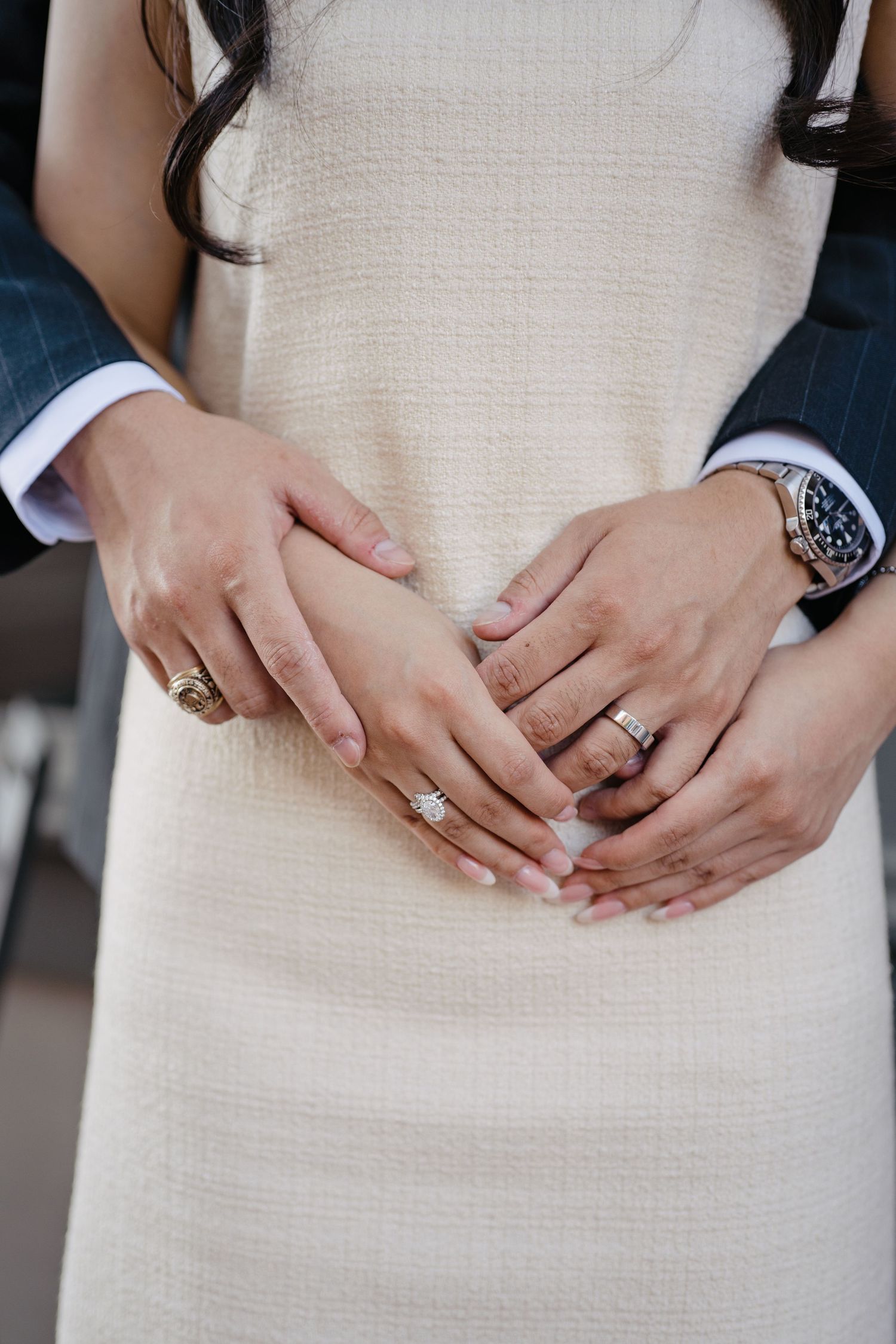 Couple holding hands showing wedding rings, bride in cream textured dress, groom in dark suit.