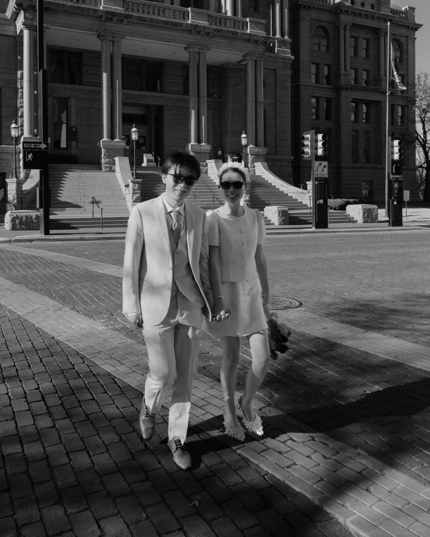 Stylish couple in coordinating white outfits walks along a brick plaza in front of a grand historic building.