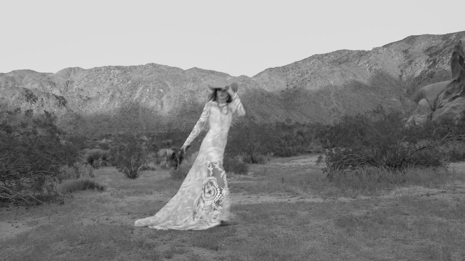 Bride in lace wedding gown poses in desert landscape with mountains in black and white photo.