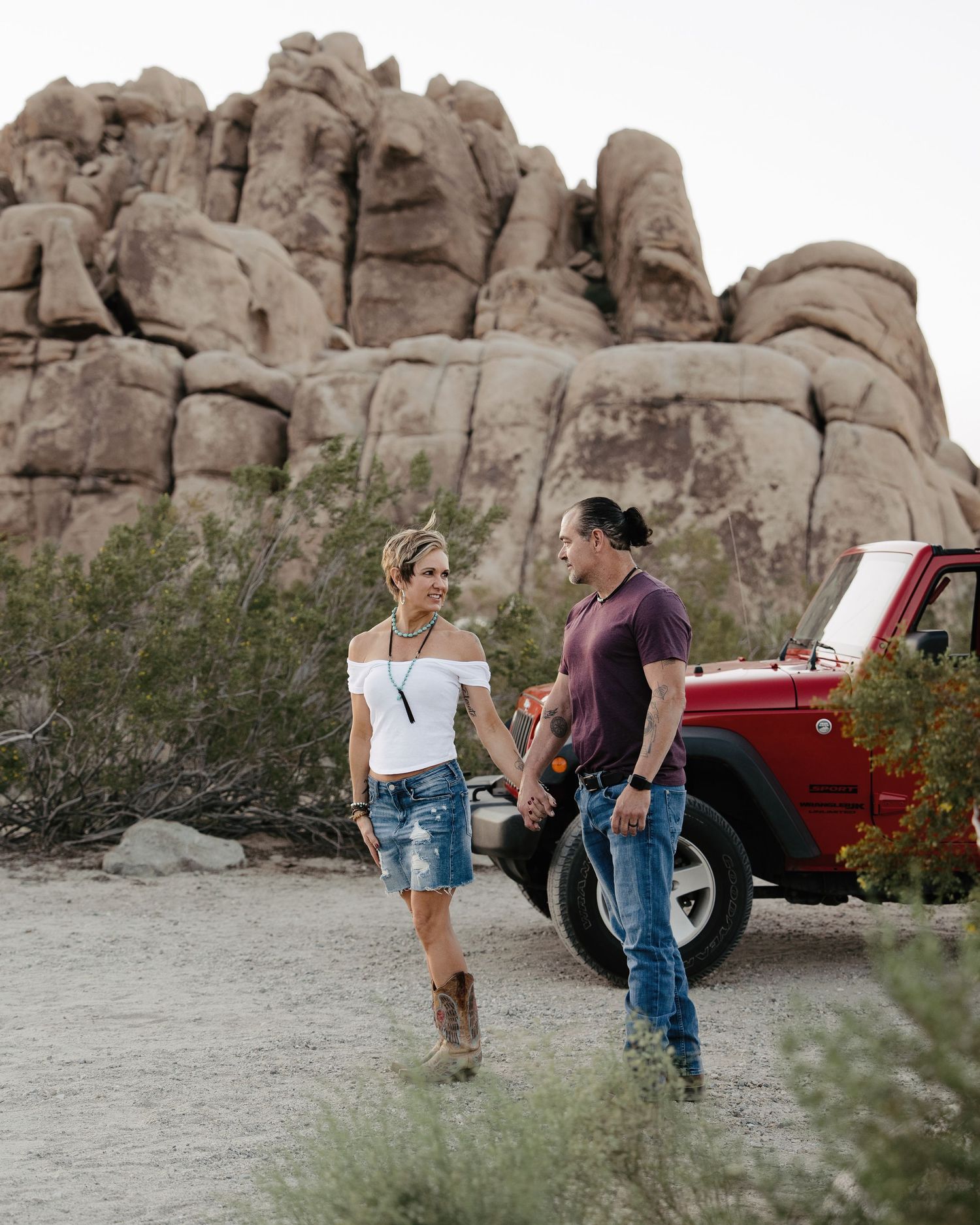 Couple holding hands and walking near a red Jeep Wrangler with large rock formations behind.