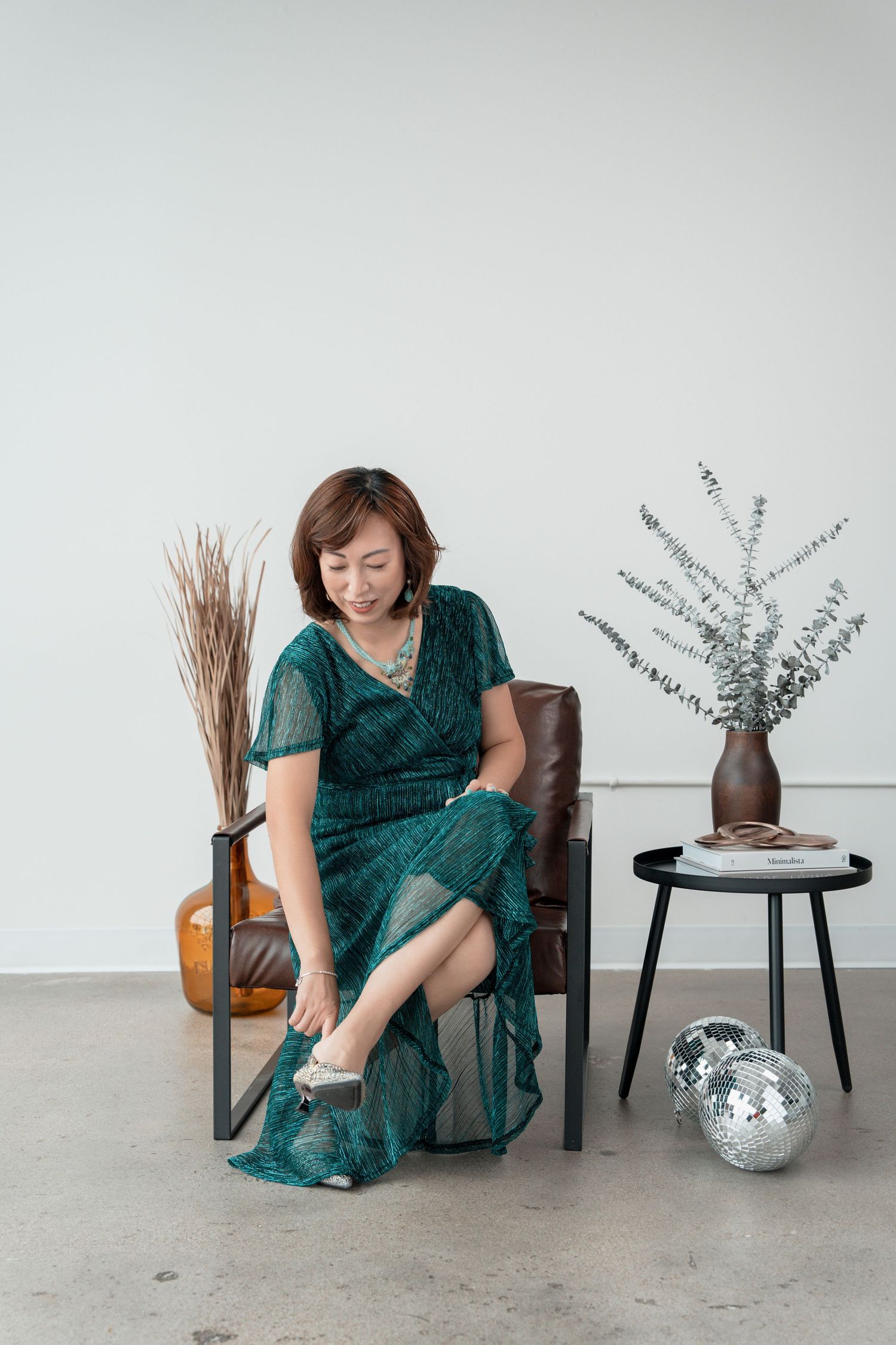 Woman in elegant green wrap dress adjusting her silver heels while seated in a leather chair.