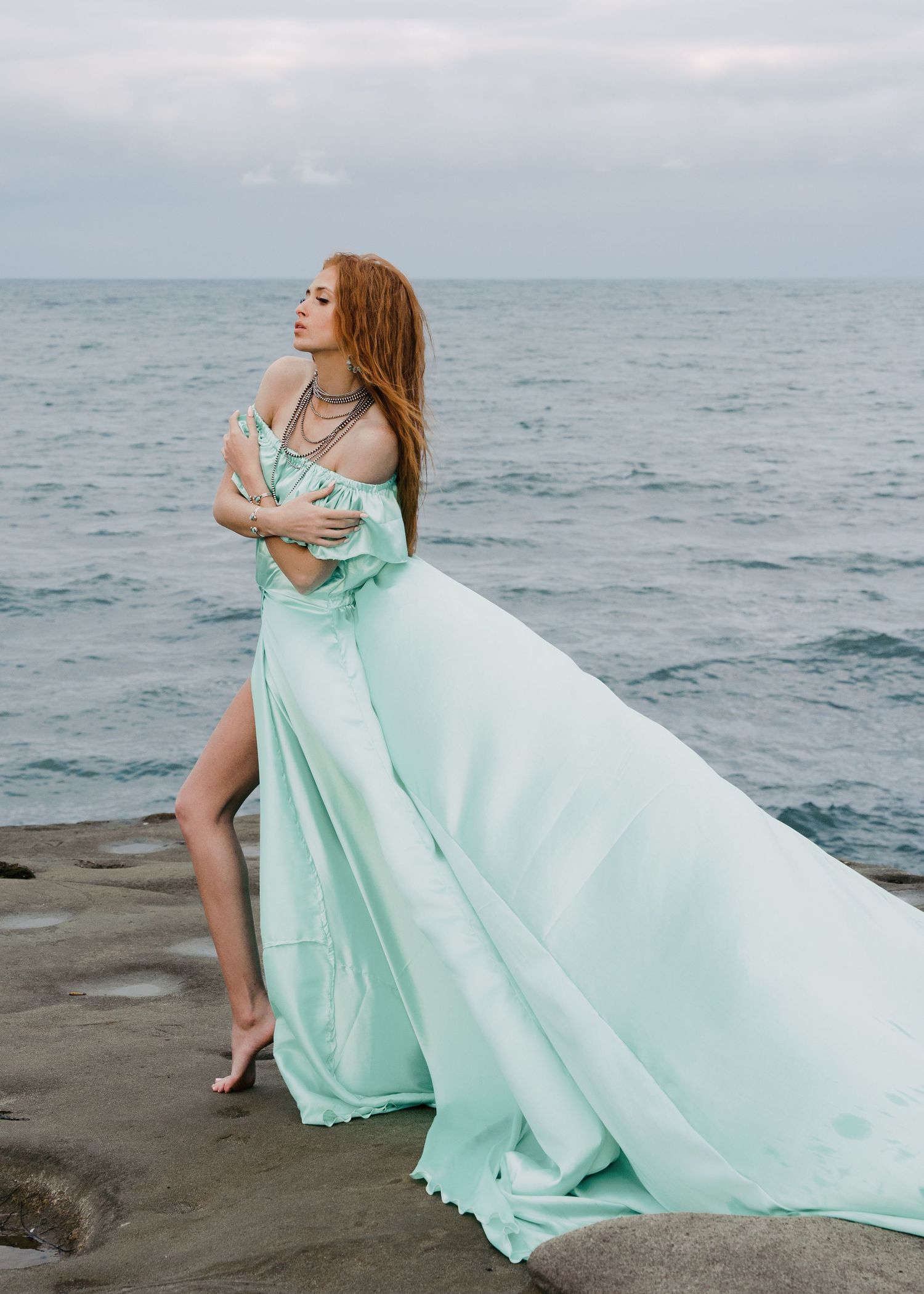 Redhead woman in mint satin off-shoulder gown with high slit posing on rocky seaside.