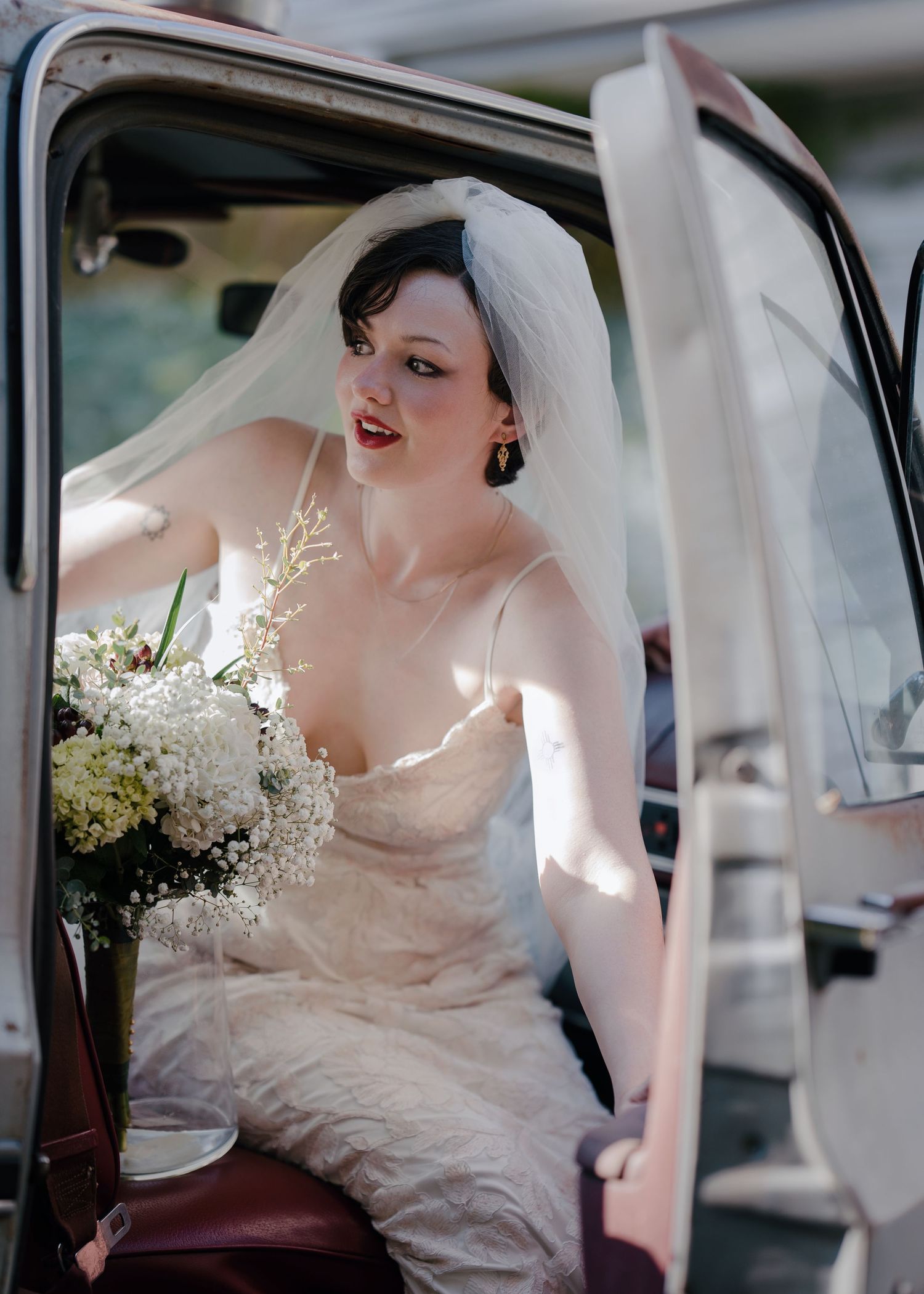 Elegant bride in lace gown holding white bouquet while sitting in vintage truck door wearing veil.
