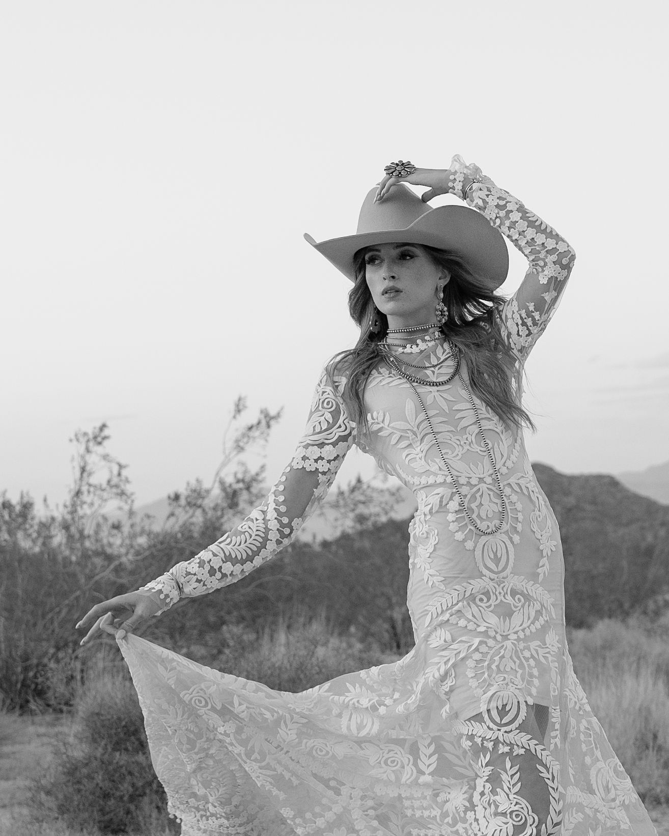 Black and white portrait of bride in lace dress and cowboy hat posing dramatically in desert setting.