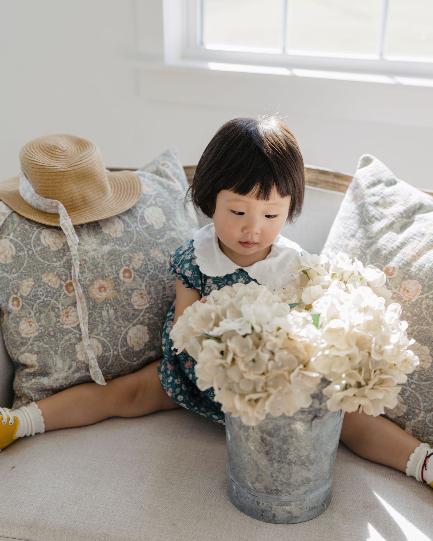 Sweet toddler girl in floral dress sitting with pillows admiring white hydrangeas in bucket.