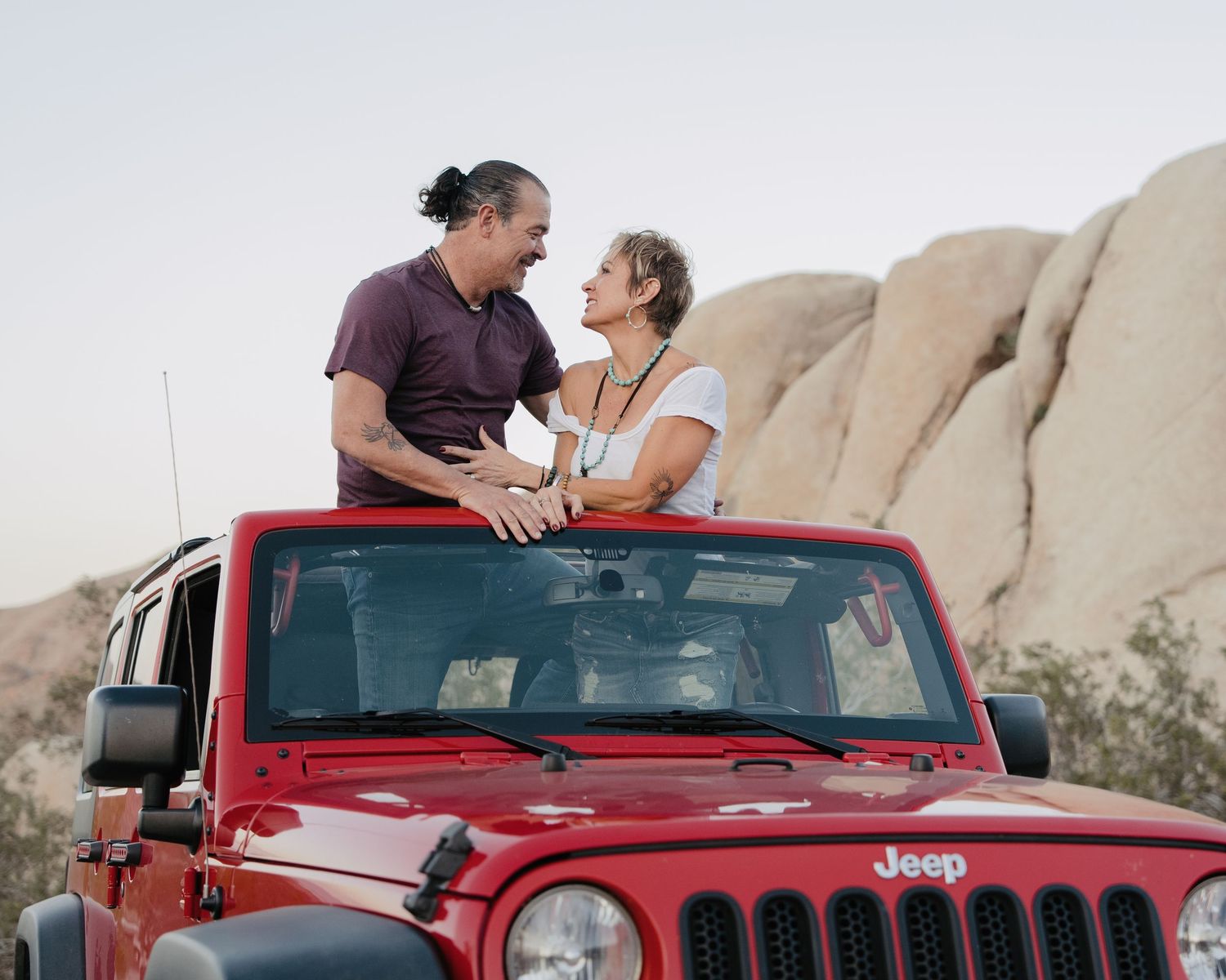 Happy couple standing through the sunroof of a red Jeep Wrangler near rocky desert boulders.