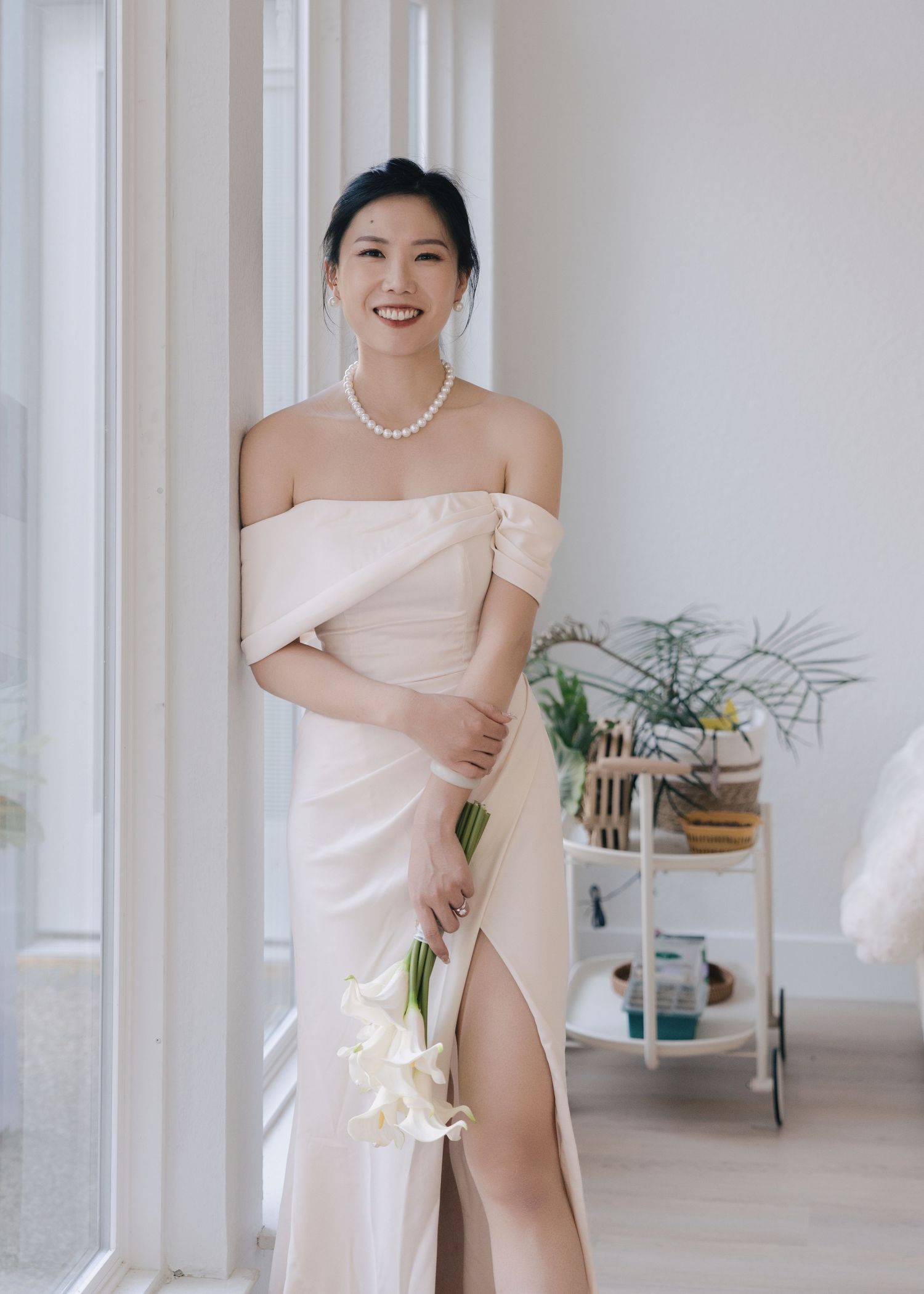 Smiling bride in ivory off-shoulder gown holds white calla lily bouquet while standing by a bright window.
