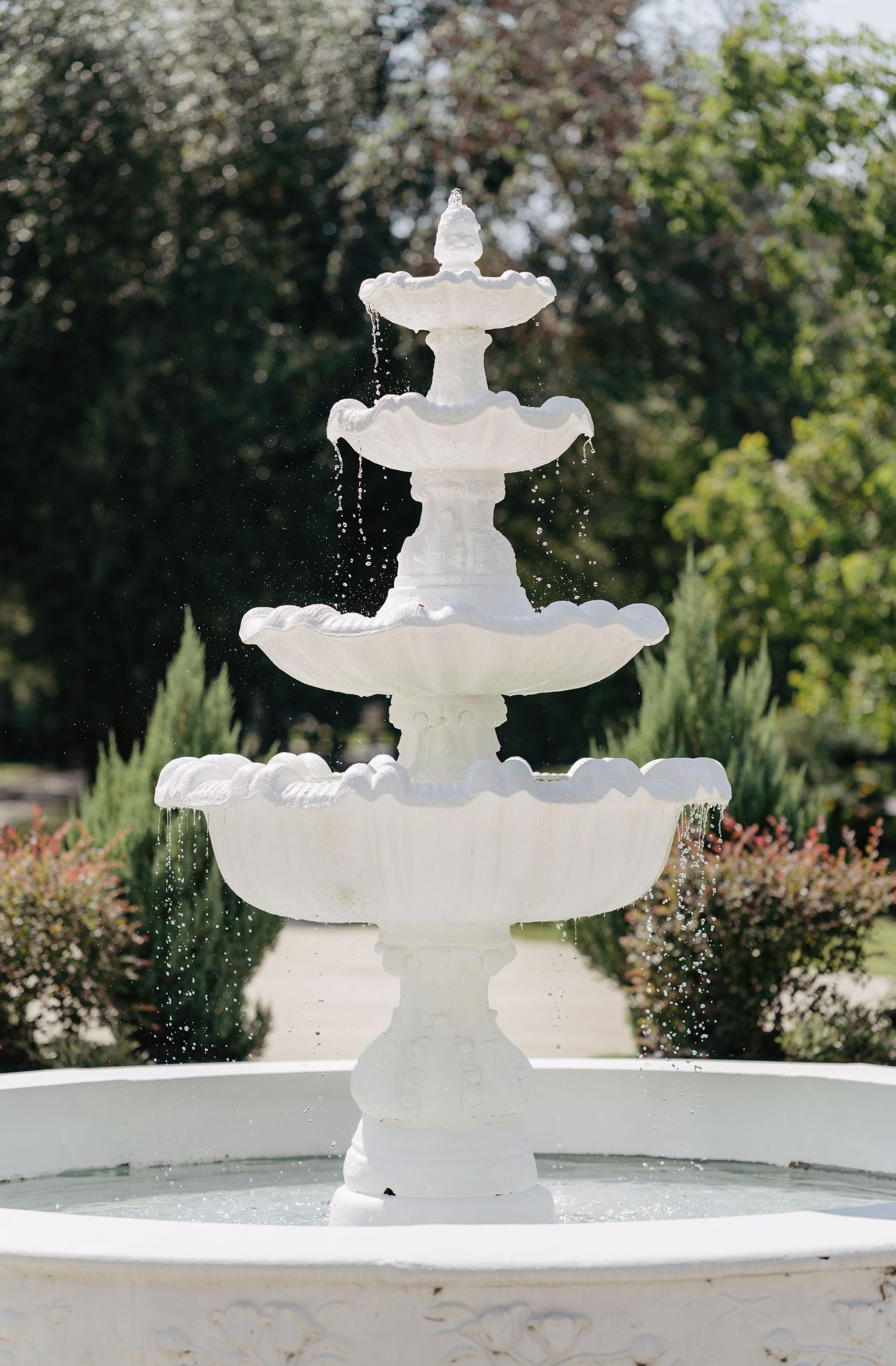 Elegant three-tiered white marble wedding fountain with cascading water in lush garden setting.