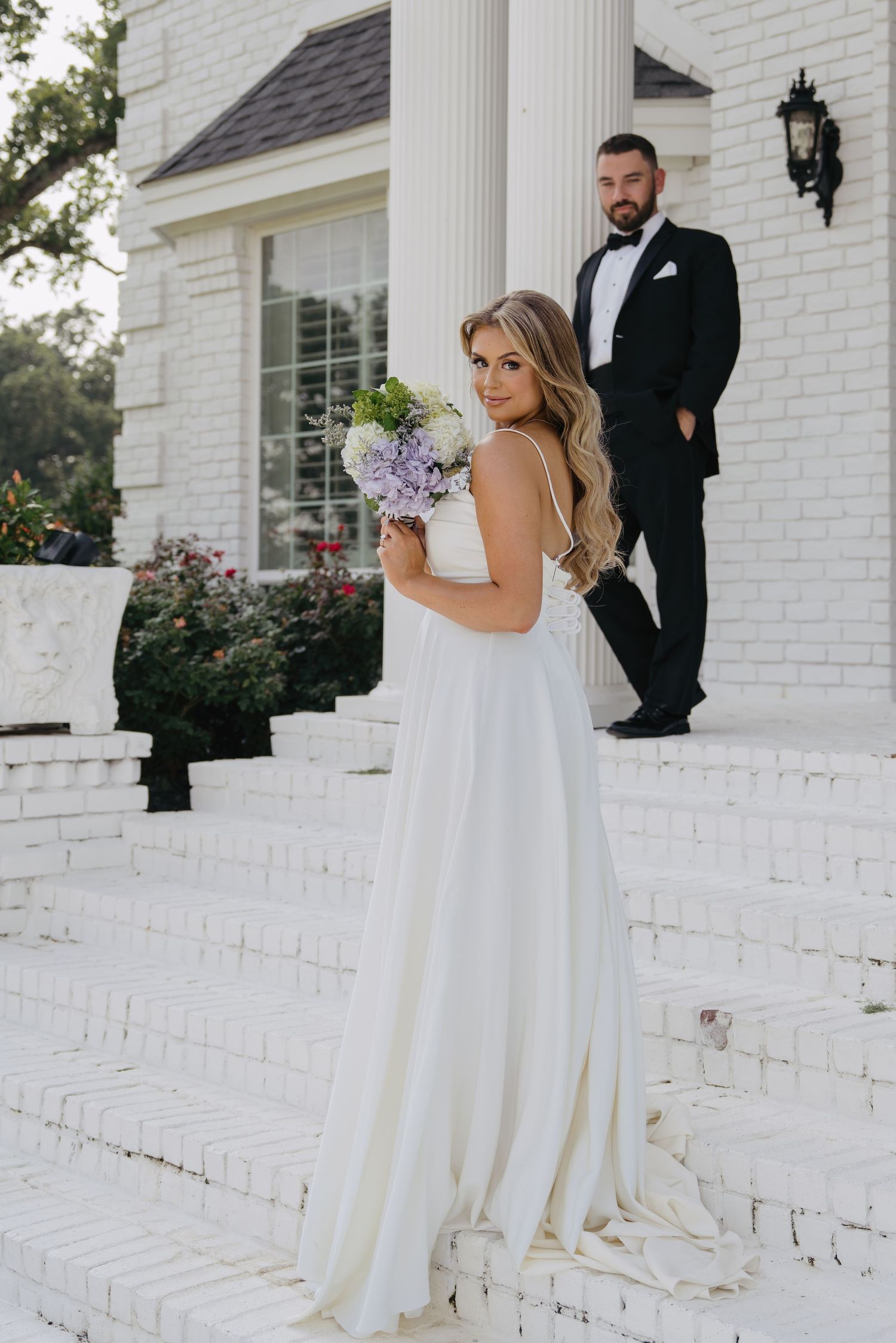 Bride in white gown holding purple bouquet poses on white brick steps with groom in tuxedo.