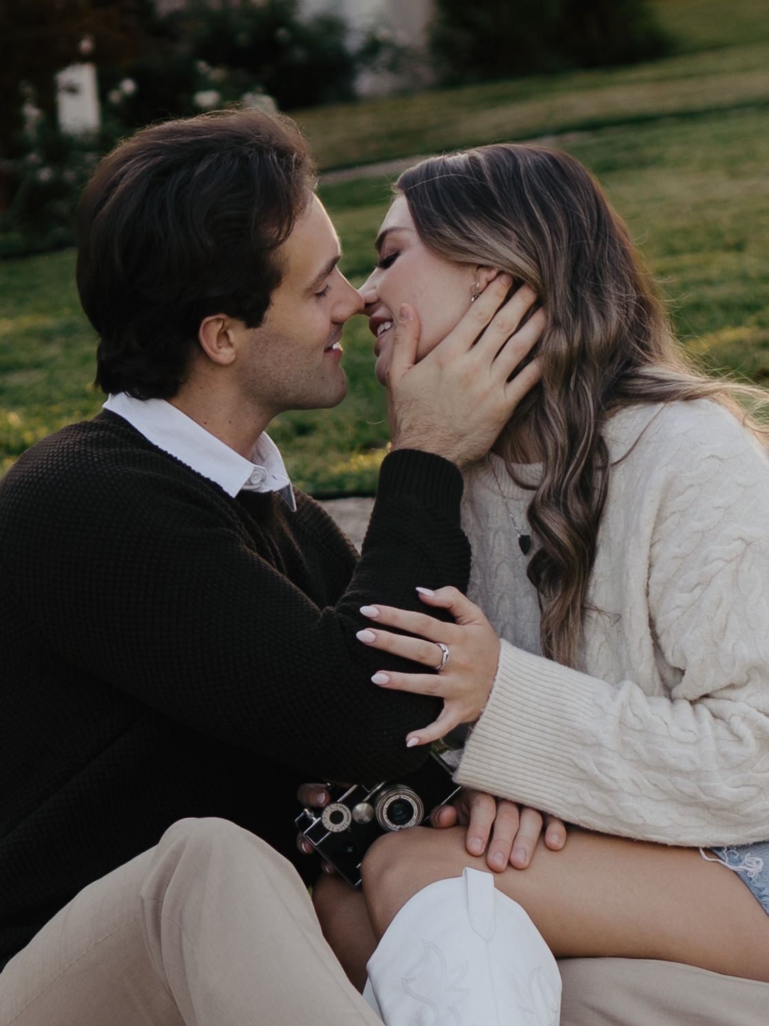 Couple sharing a romantic moment outdoors, woman in cream sweater smiling as man gently holds her face near green grass.
