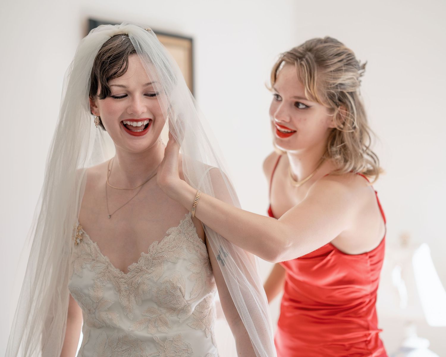 Joyful bride in lace gown and veil smiling as bridesmaid in red dress adjusts her veil.
